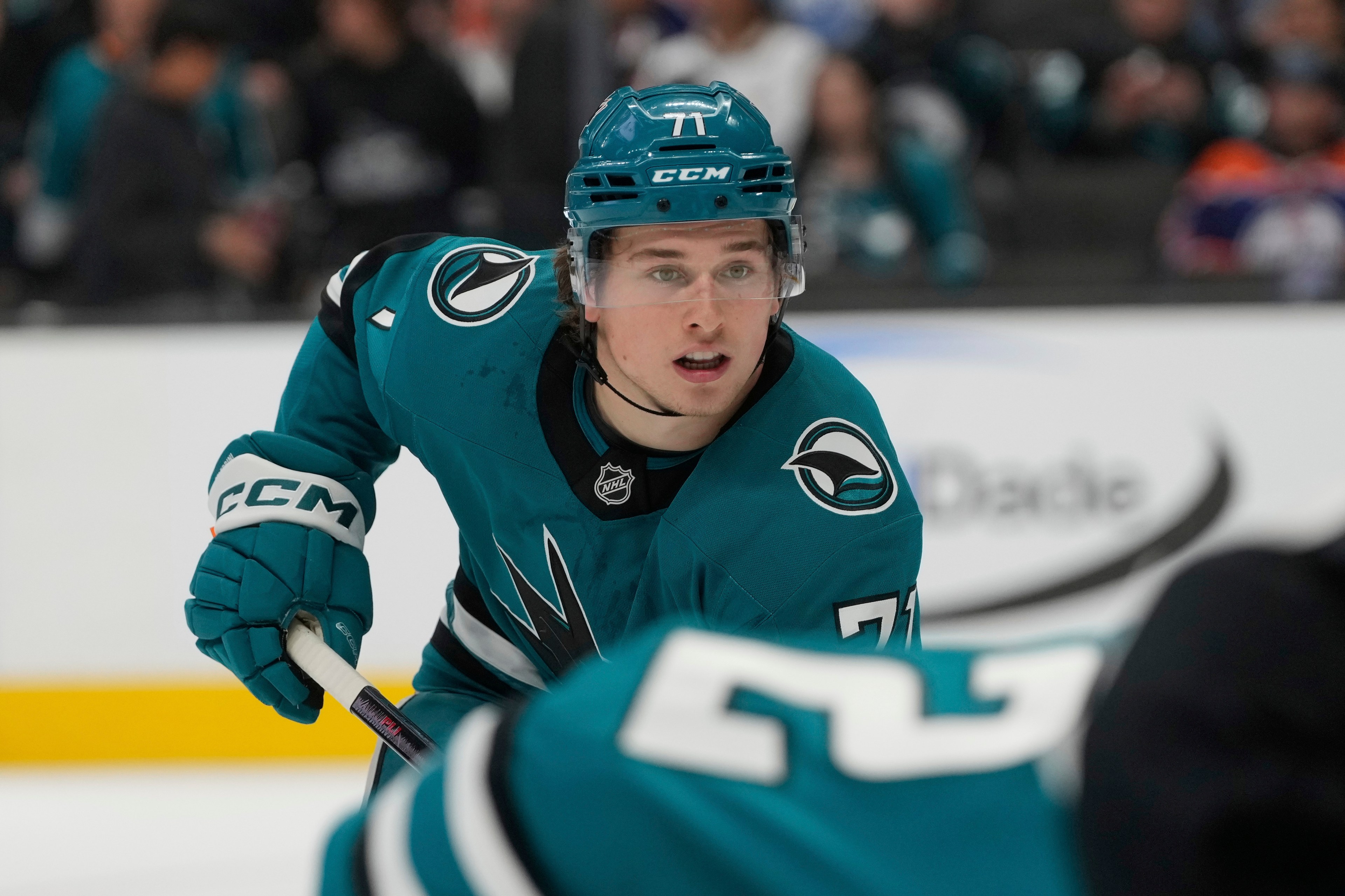 A hockey player wearing a teal jersey and helmet skates on the ice, gripping a stick. Spectators blur in the background.