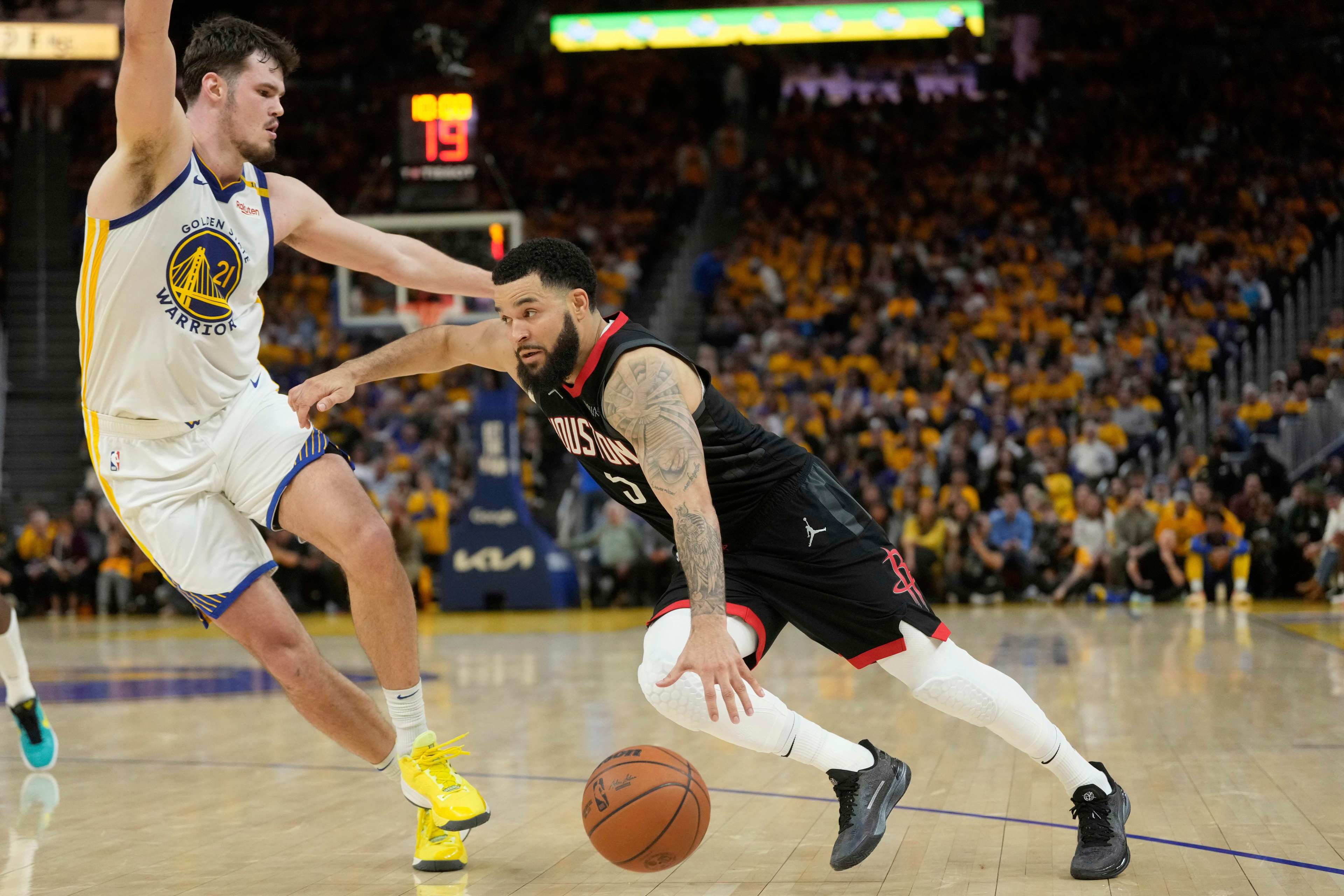 A basketball player in a black uniform dribbles past a defending player in a white and yellow uniform on a court, with a crowd in the background.