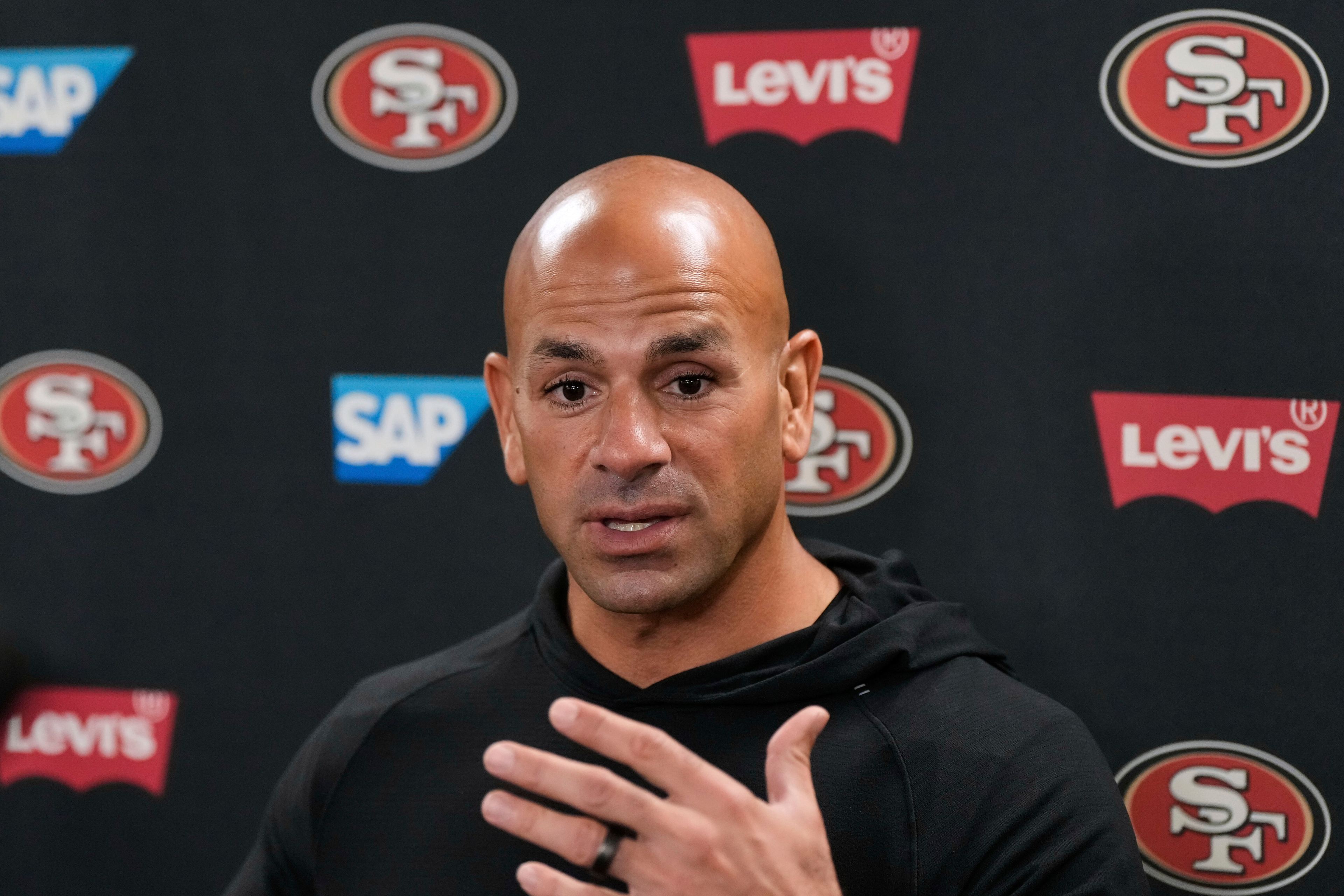 A bald man in a black hoodie speaks, gesturing with his hand. Behind him are logos for the San Francisco 49ers, SAP, and Levi's on a black background.