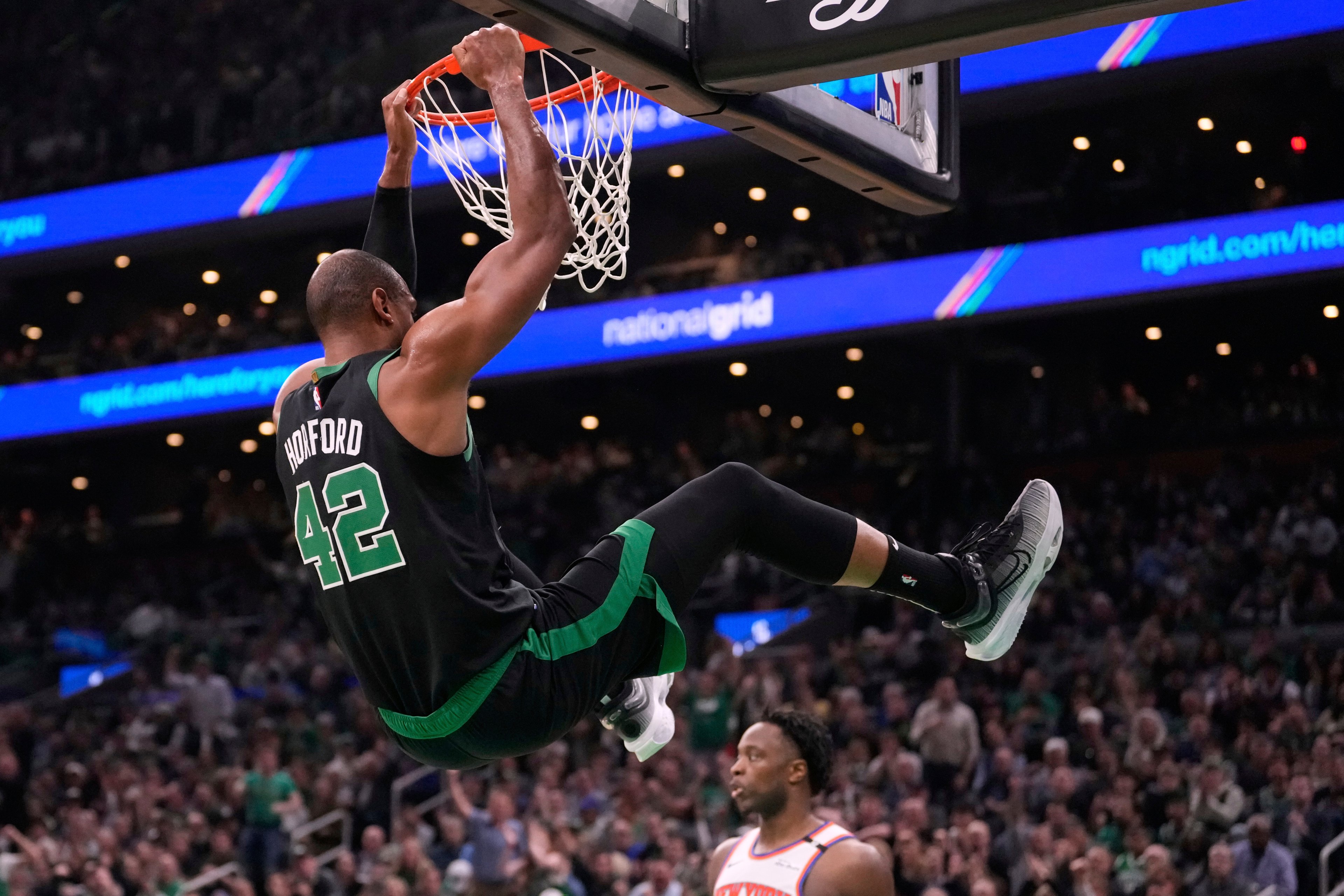 A basketball player in a green jersey labeled &quot;42&quot; hangs from the rim after dunking. Another player in a white jersey watches, with a crowded arena in the background.