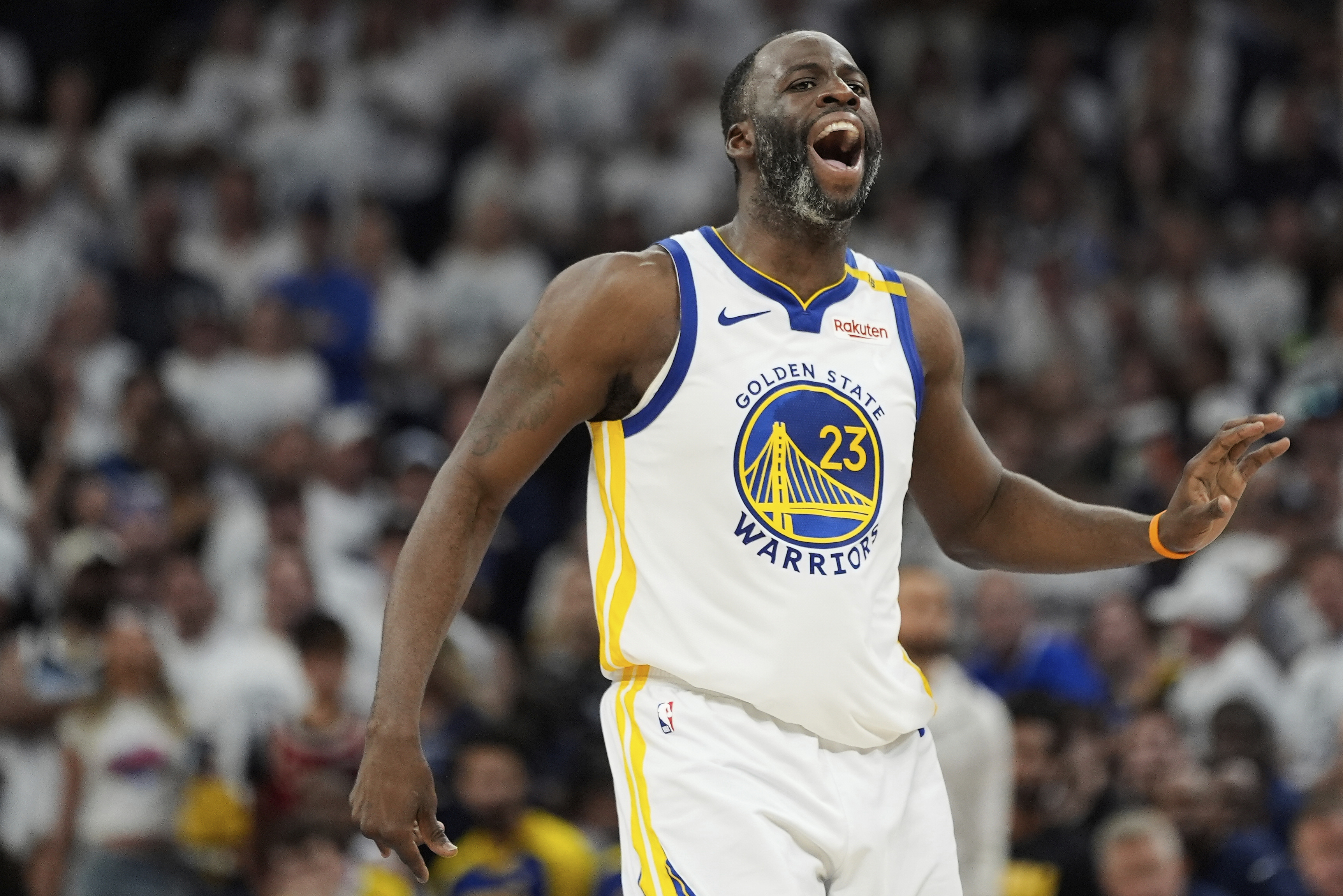 A basketball player in a Golden State Warriors jersey passionately shouts during a game, with a blurred crowd in the background.