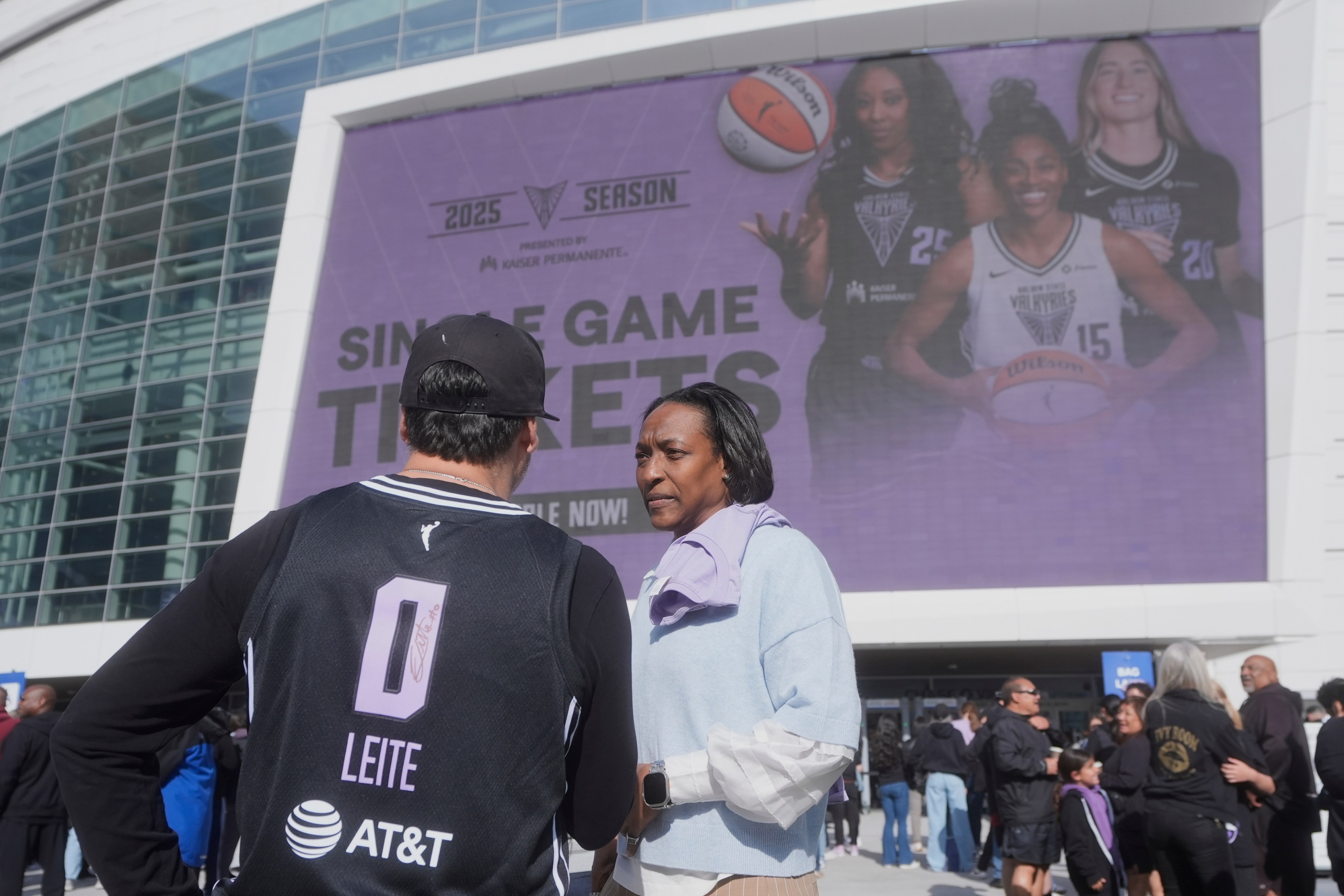 A group of people stands outside a building with a large poster advertising 2025 season tickets for a women's basketball team, featuring three players.