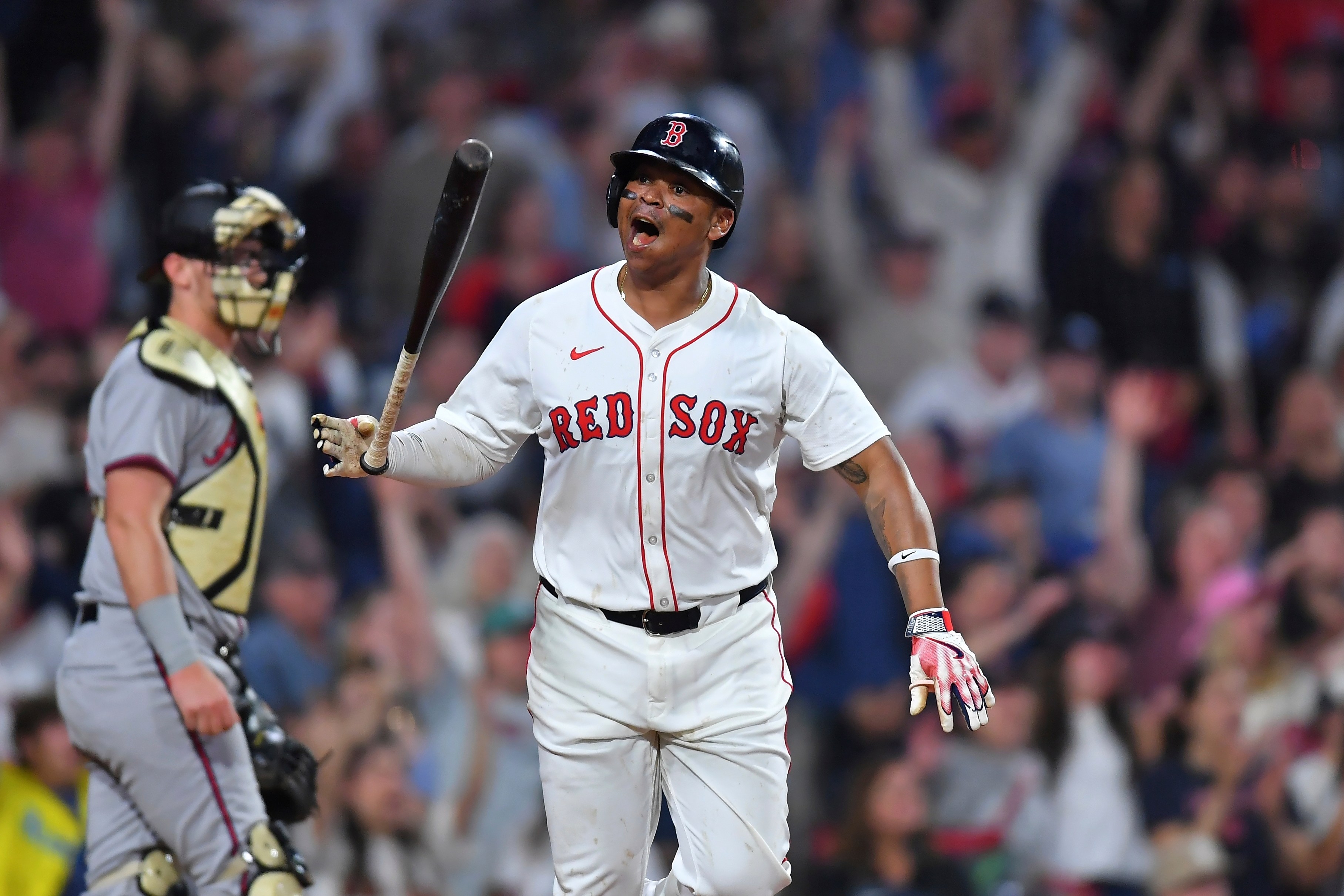 A baseball player in a Red Sox uniform stands excitedly on the field, holding a bat. A catcher is nearby, and the crowd is visible in the background.