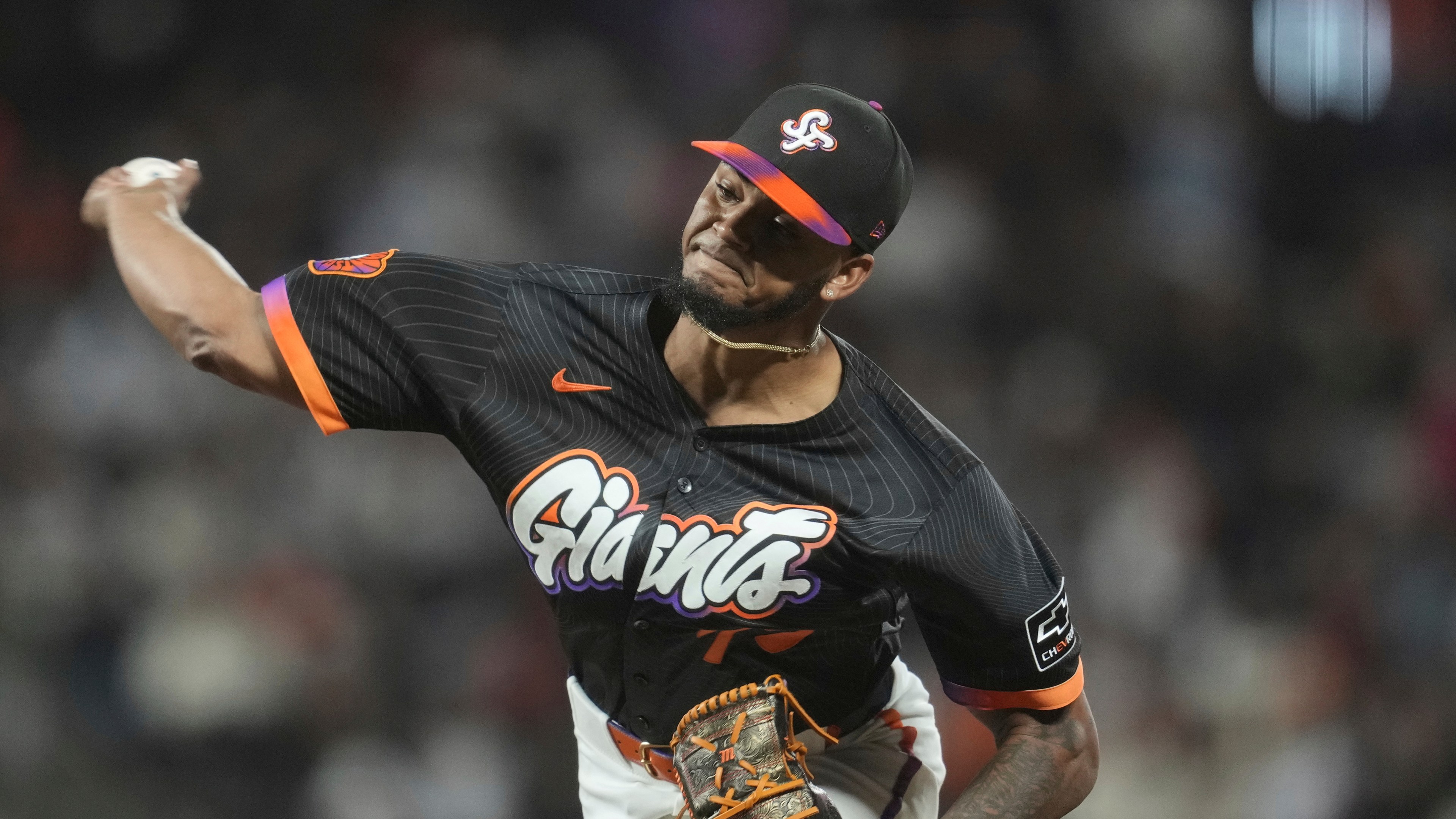 A baseball player in a black &quot;Giants&quot; jersey and cap is pitching. The jersey has orange and purple accents, and he wears a glove with matching colors.