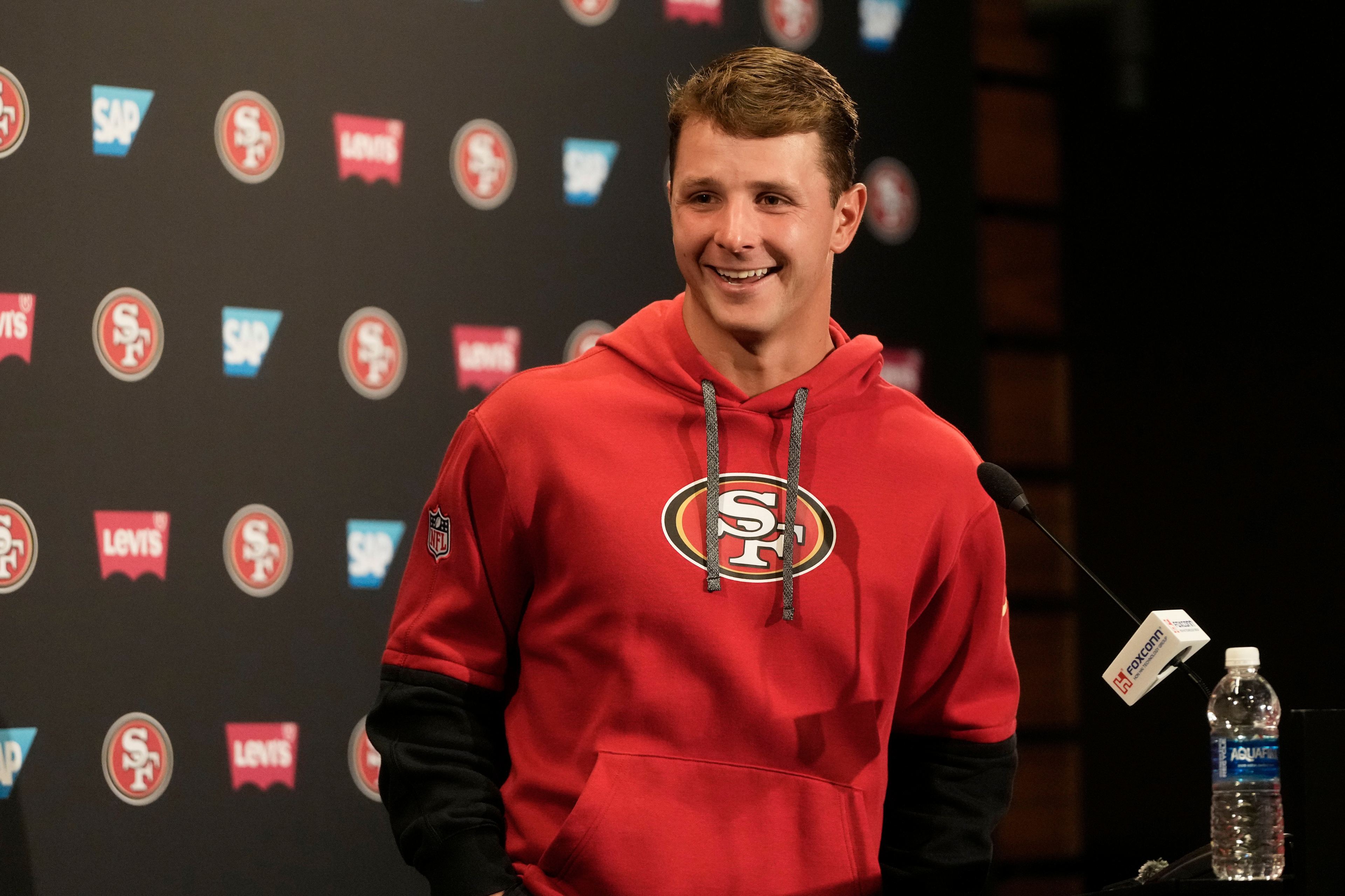 A smiling man in a red 49ers hoodie stands at a podium with microphones, in front of a branded backdrop featuring SAP and Levi's logos.