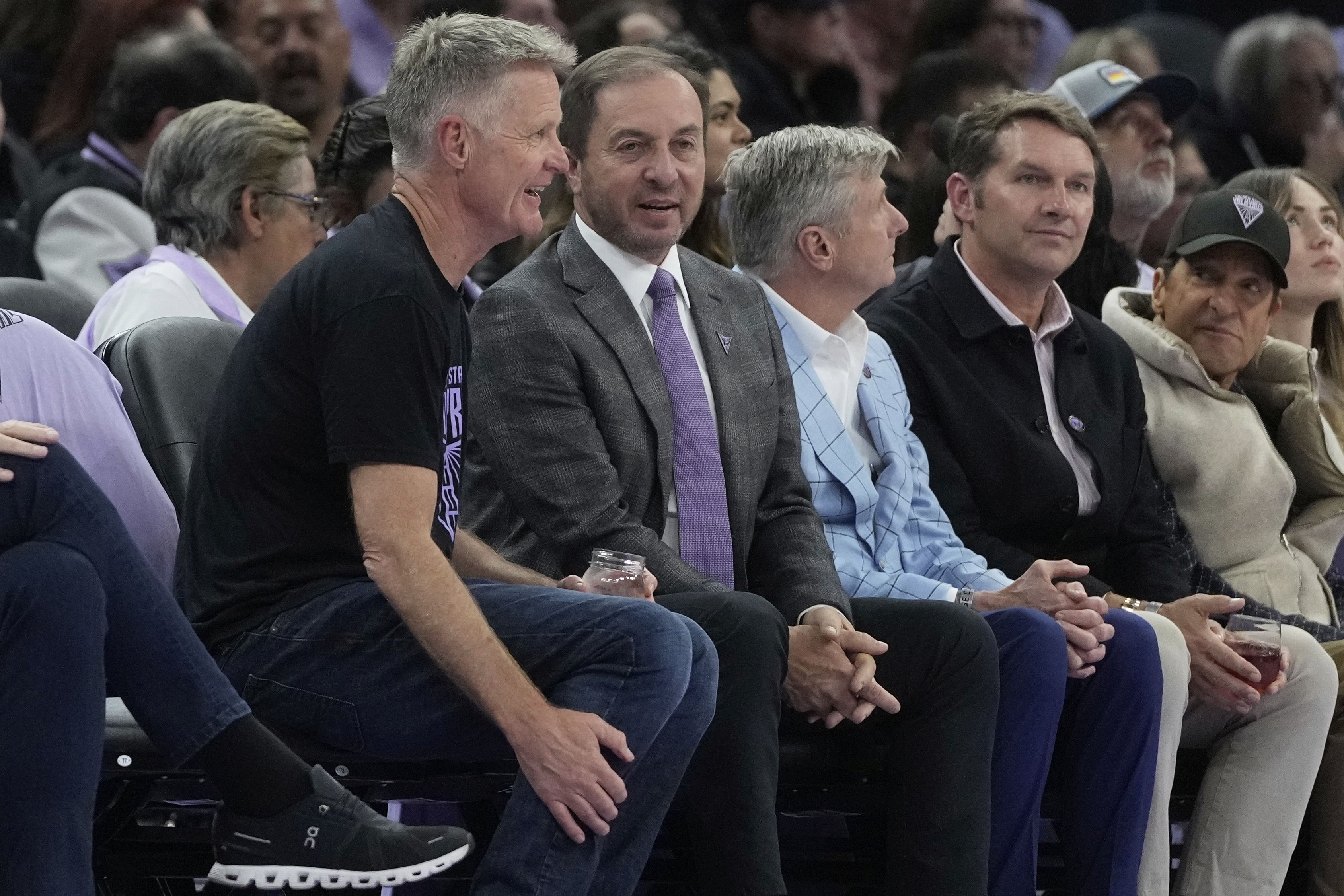 A group of men are sitting in a row at an event. One man wears a black t-shirt and jeans, laughing. Others are in suits and jackets, engaged in conversation.