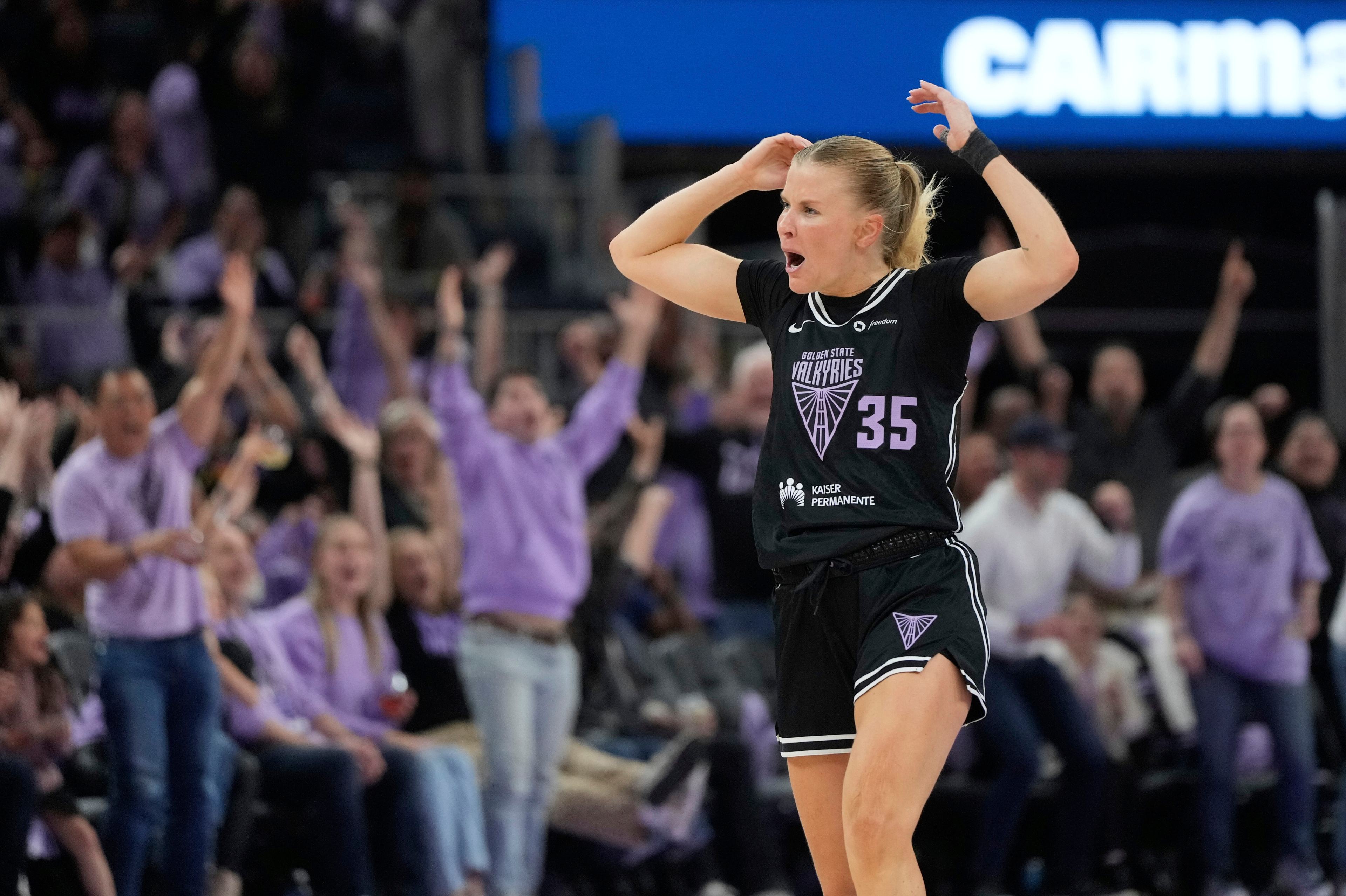 A basketball player in a black uniform gestures with excitement on the court. The crowd in the background cheers with raised arms, creating an energetic atmosphere.