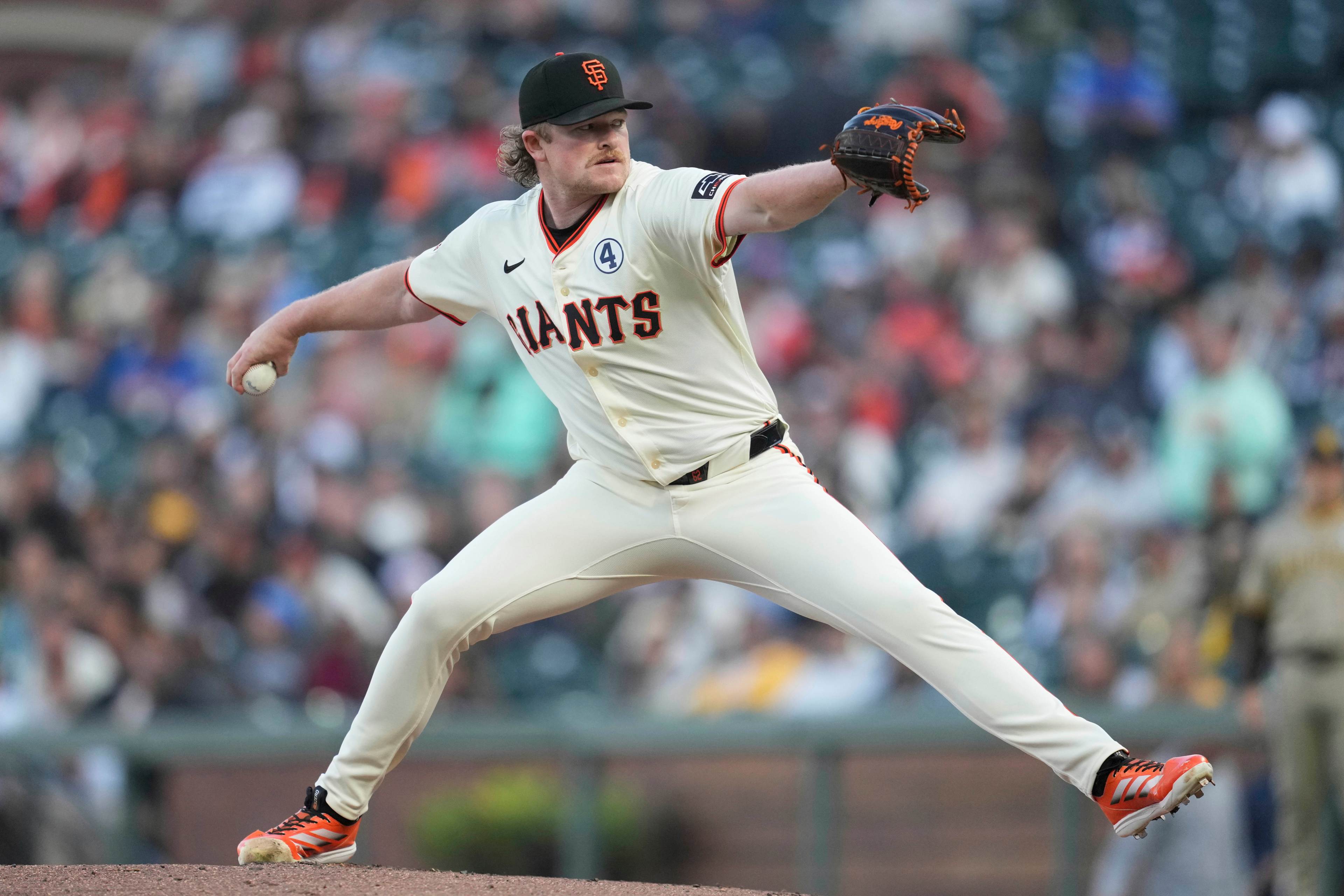 A baseball player in a white &quot;Giants&quot; uniform is pitching on the mound, wearing an SF cap and orange cleats, with a blurred crowd in the background.