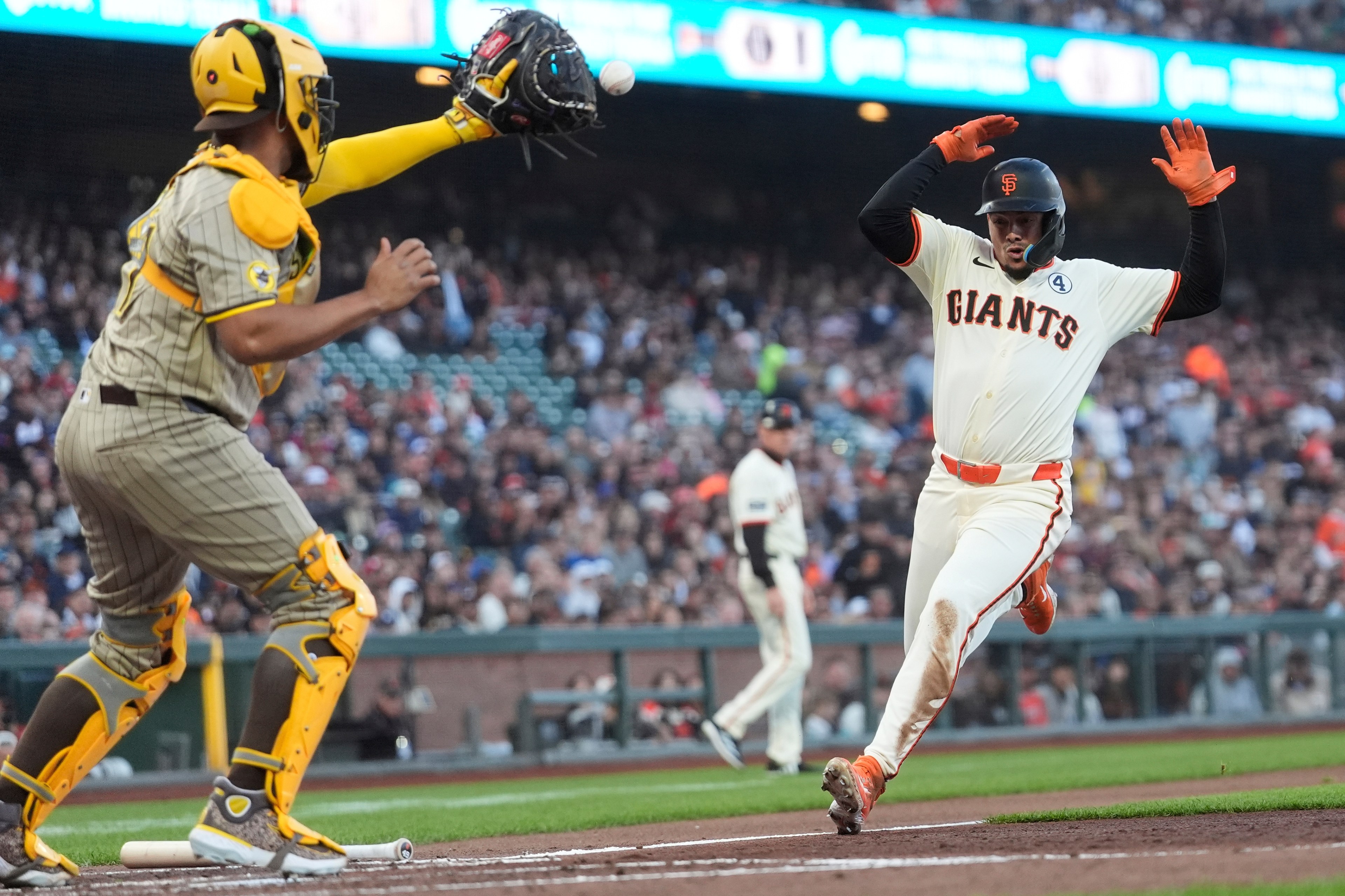 A baseball player from the Giants slides toward home plate as a catcher in yellow gear reaches with his glove to tag him, with spectators in the background.