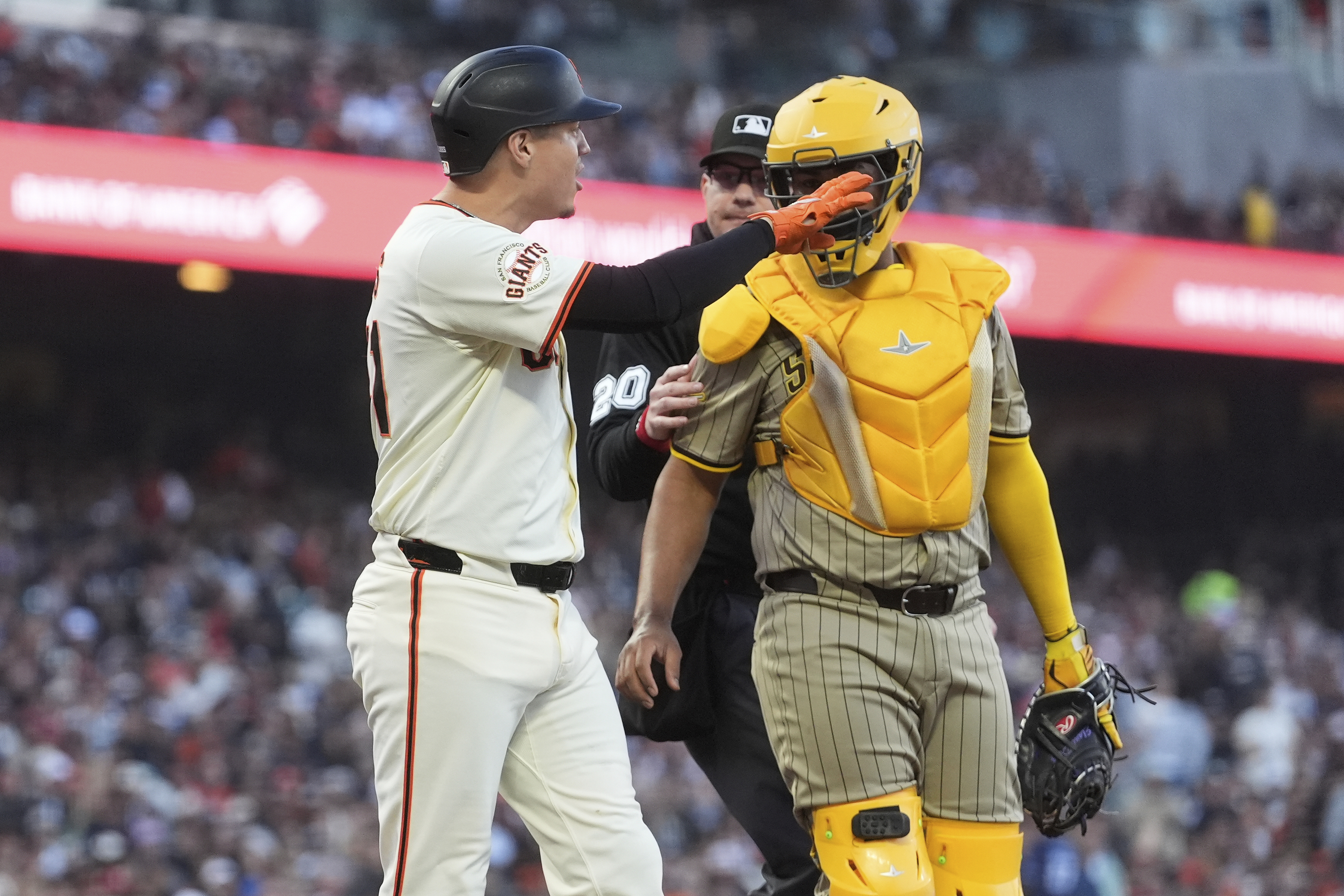 A baseball player in a white uniform gestures near a catcher in yellow protective gear. An umpire stands between them, with the crowd blurred in the background.
