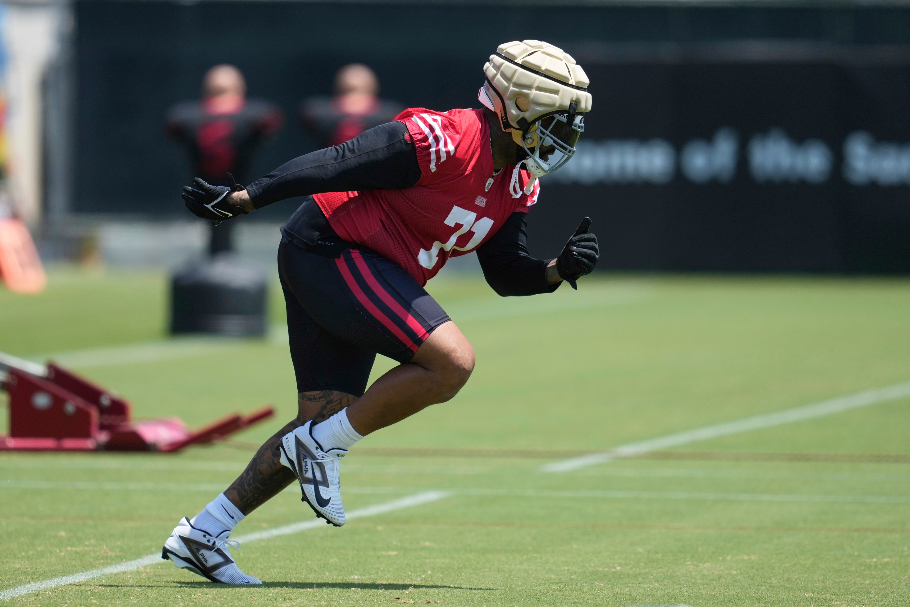 A football player wearing a padded helmet and red jersey runs on a grassy field during practice, with blurred training equipment in the background.