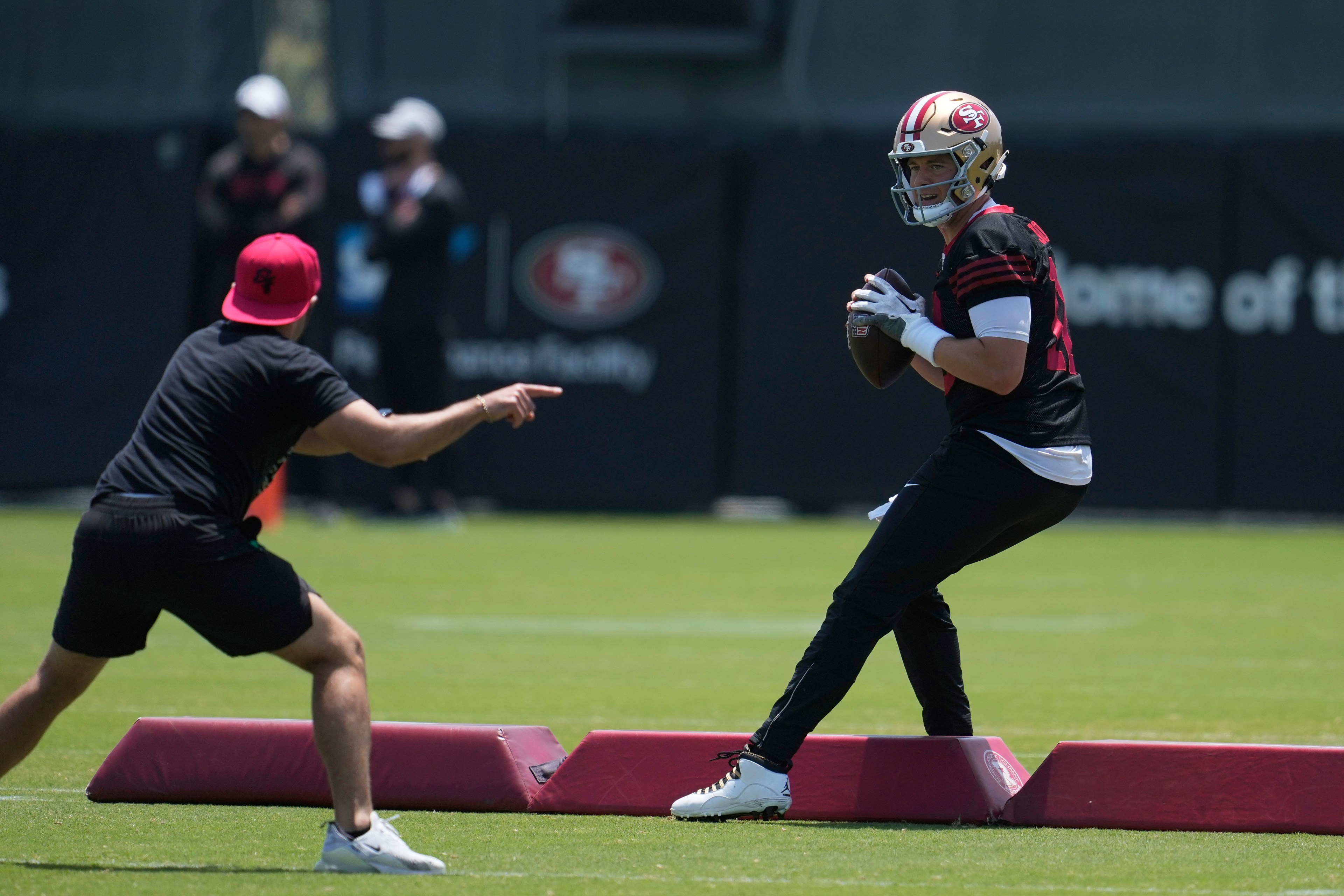 A football player in a helmet holds a ball, poised to throw. Another person in a red cap points at him during a practice session on a field.