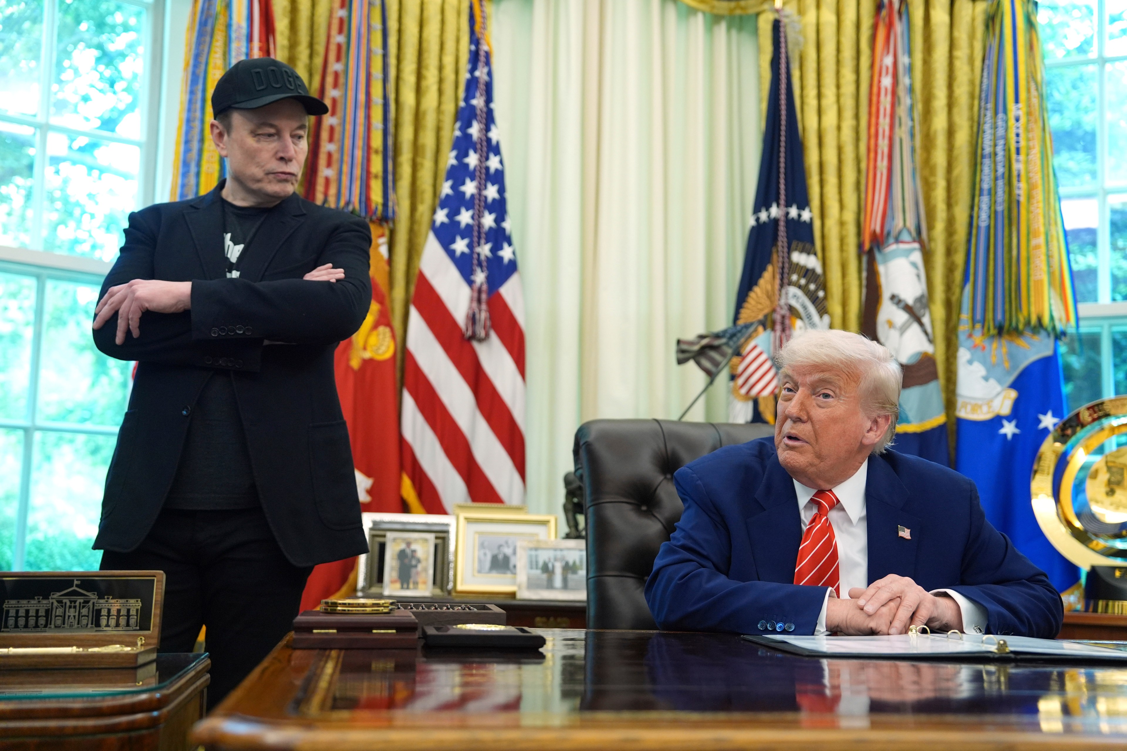 A man in a black cap and jacket stands with arms crossed near another man in a blue suit seated at a desk. Flags and ornate decorations are visible behind them.