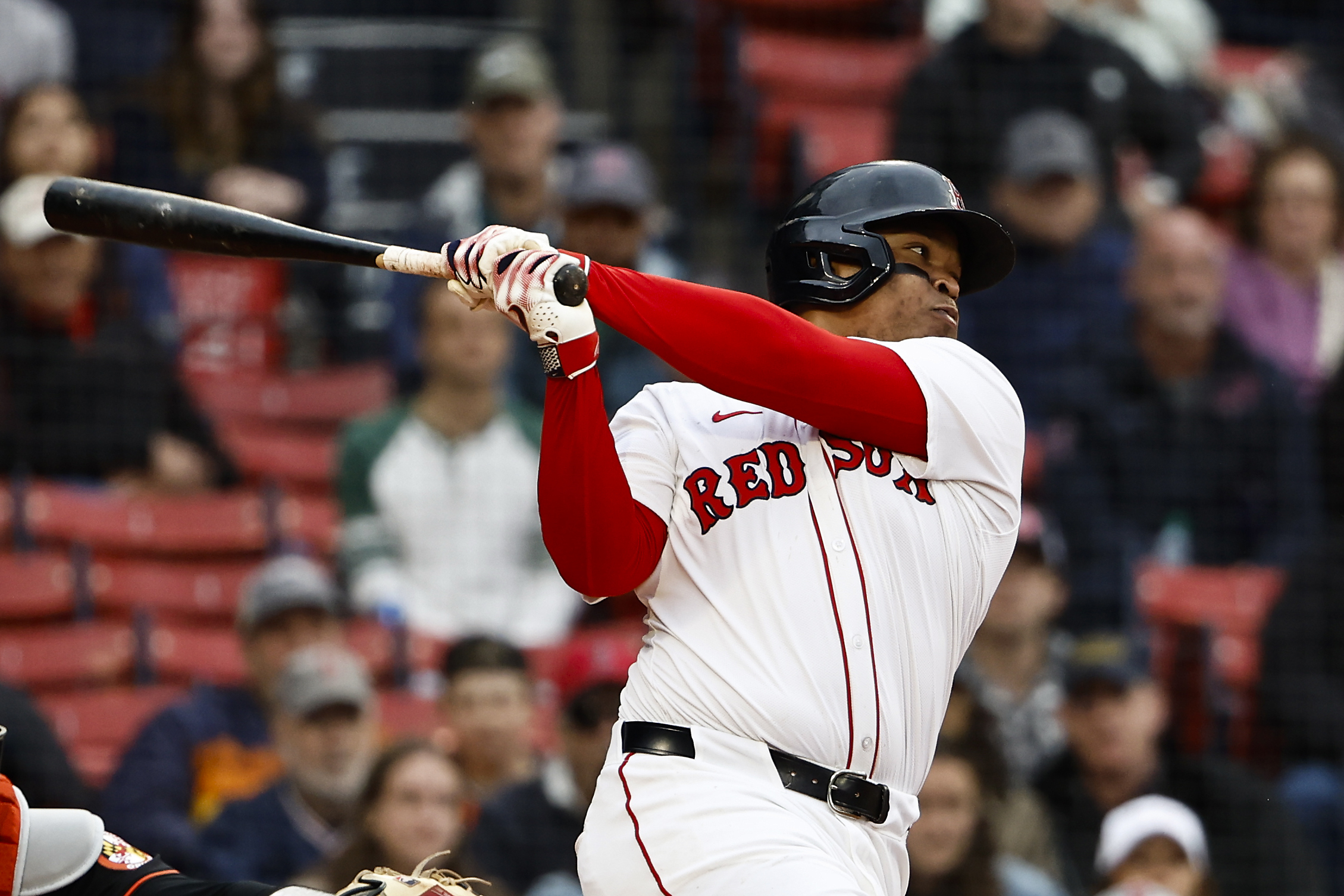 A baseball player in a white Red Sox uniform is batting. He has just swung his bat, and spectators are visible in the blurred stadium background.