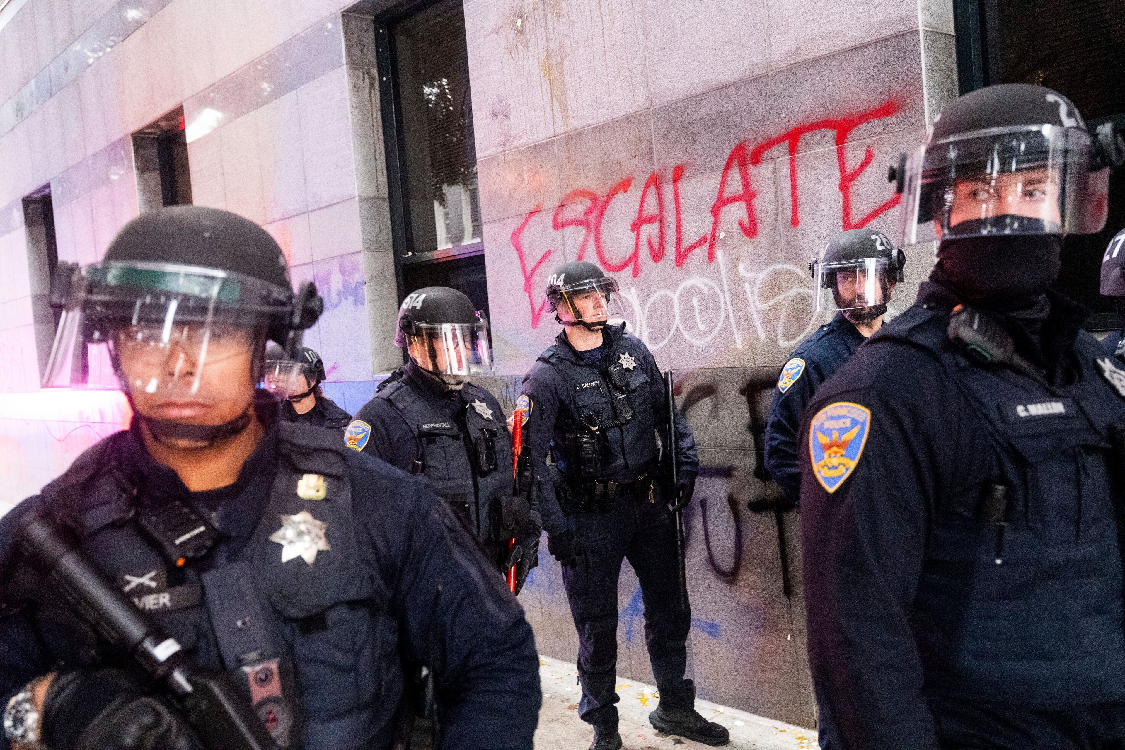 Police officers in riot gear stand in front of a building wall with the word &quot;ESCALATE&quot; spray-painted in red.