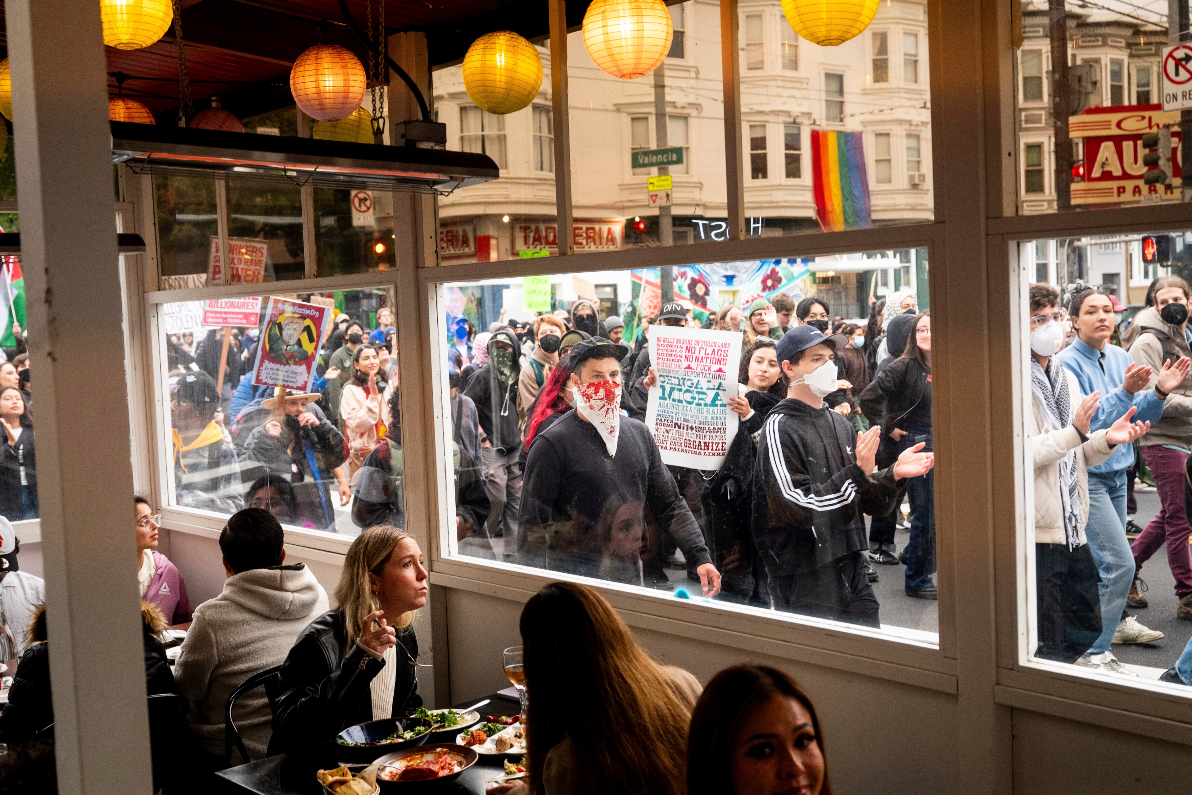 Inside a restaurant, diners eat near a window. Outside, a protest takes place with people holding signs, some wearing masks, and one rainbow flag visible.