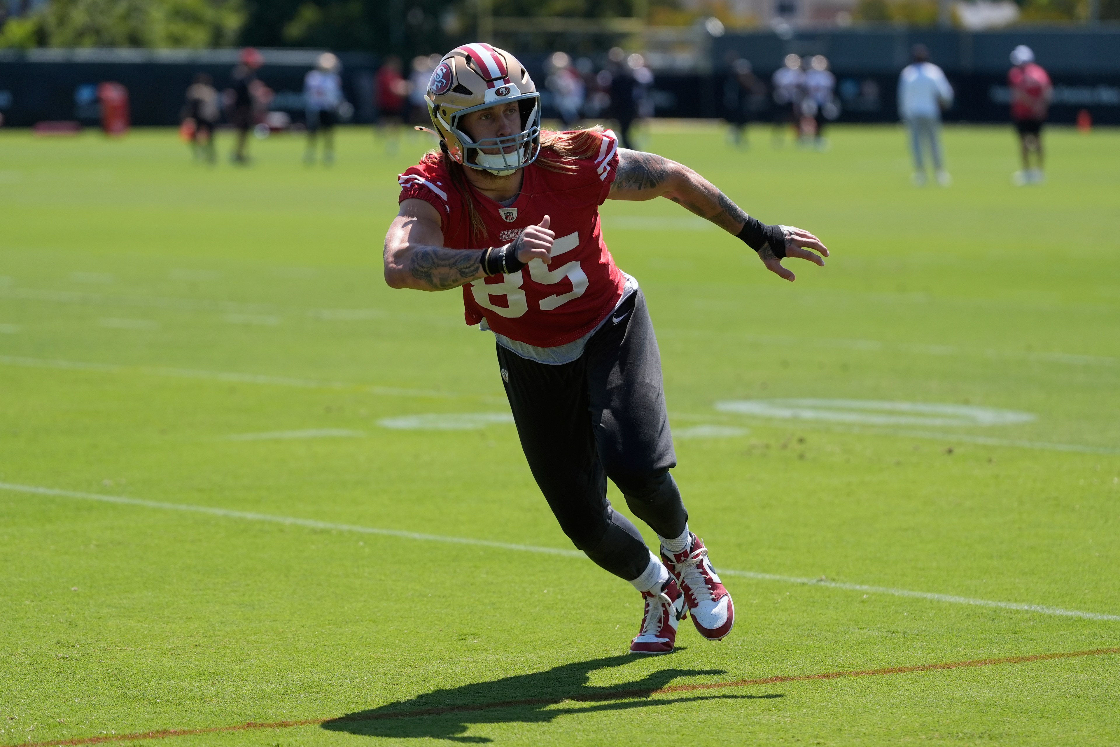 A football player in a red jersey and black pants is running on a green field during practice. He's wearing a helmet and has tattoos on his arms.