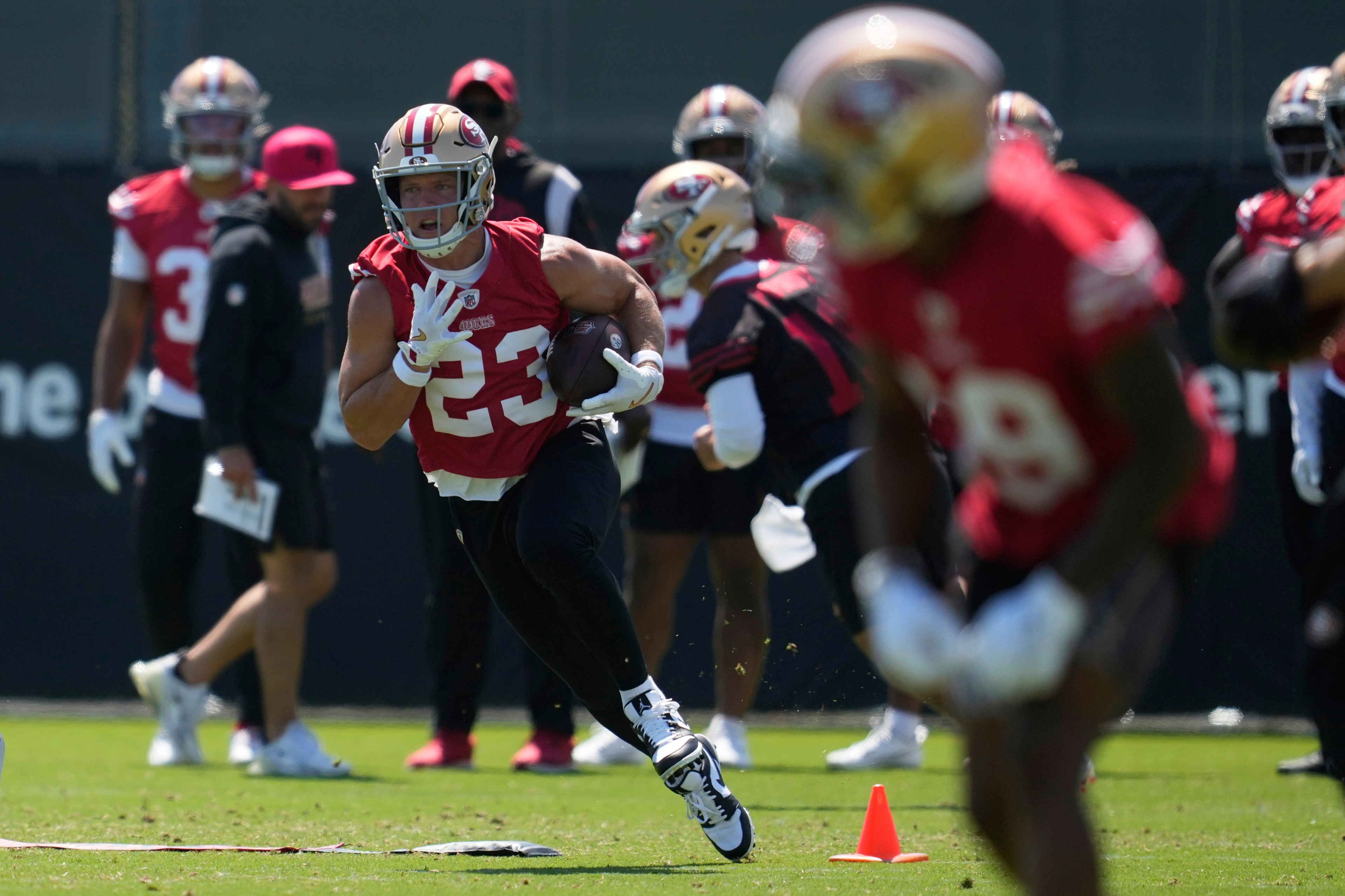 A football player in red uniform and helmet, holding a ball, is running on a green field. Other players and coaches in red are visible in the background.