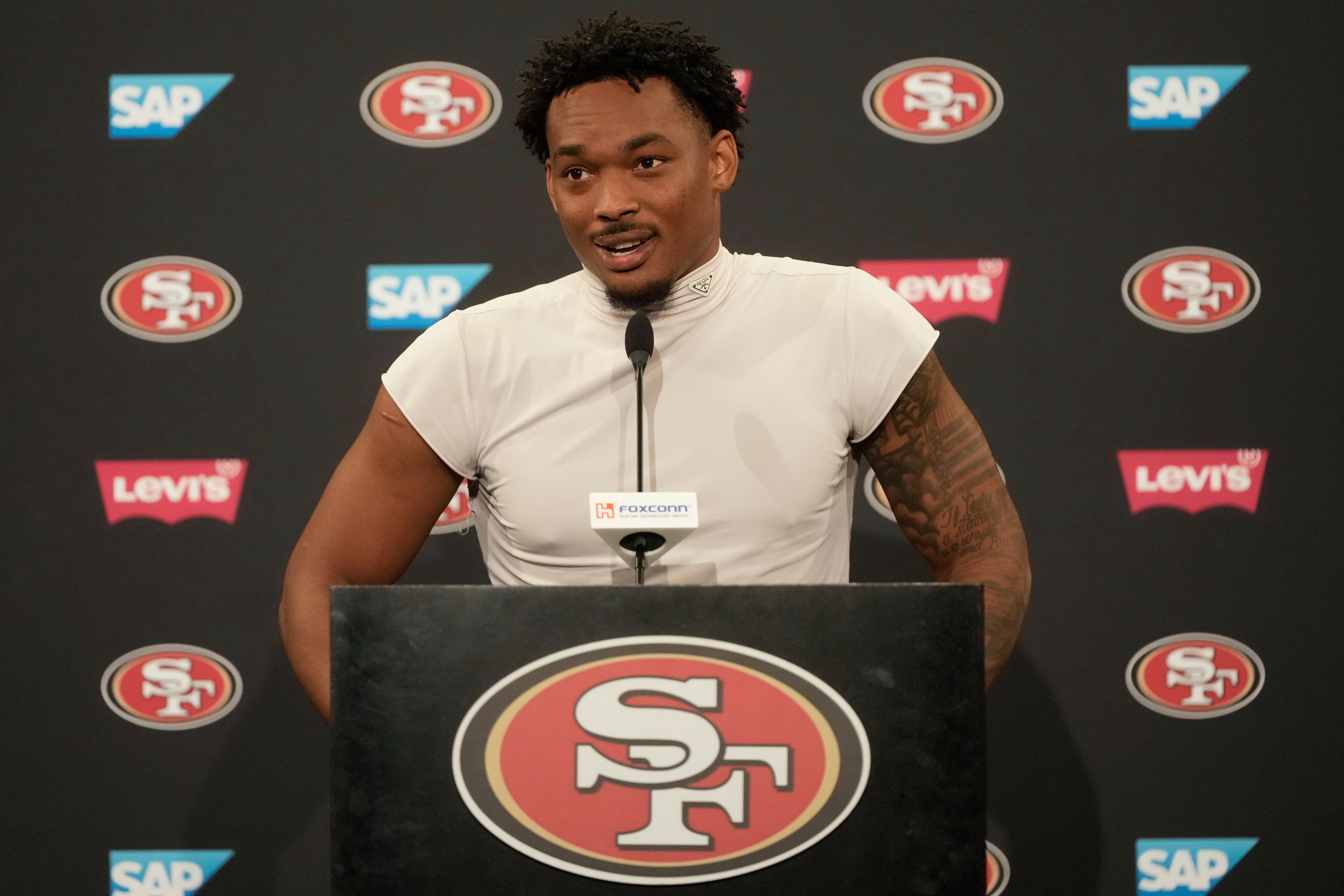 A man in a white sleeveless shirt speaks at a podium with a 49ers logo. The background features logos for SAP and Levi's.
