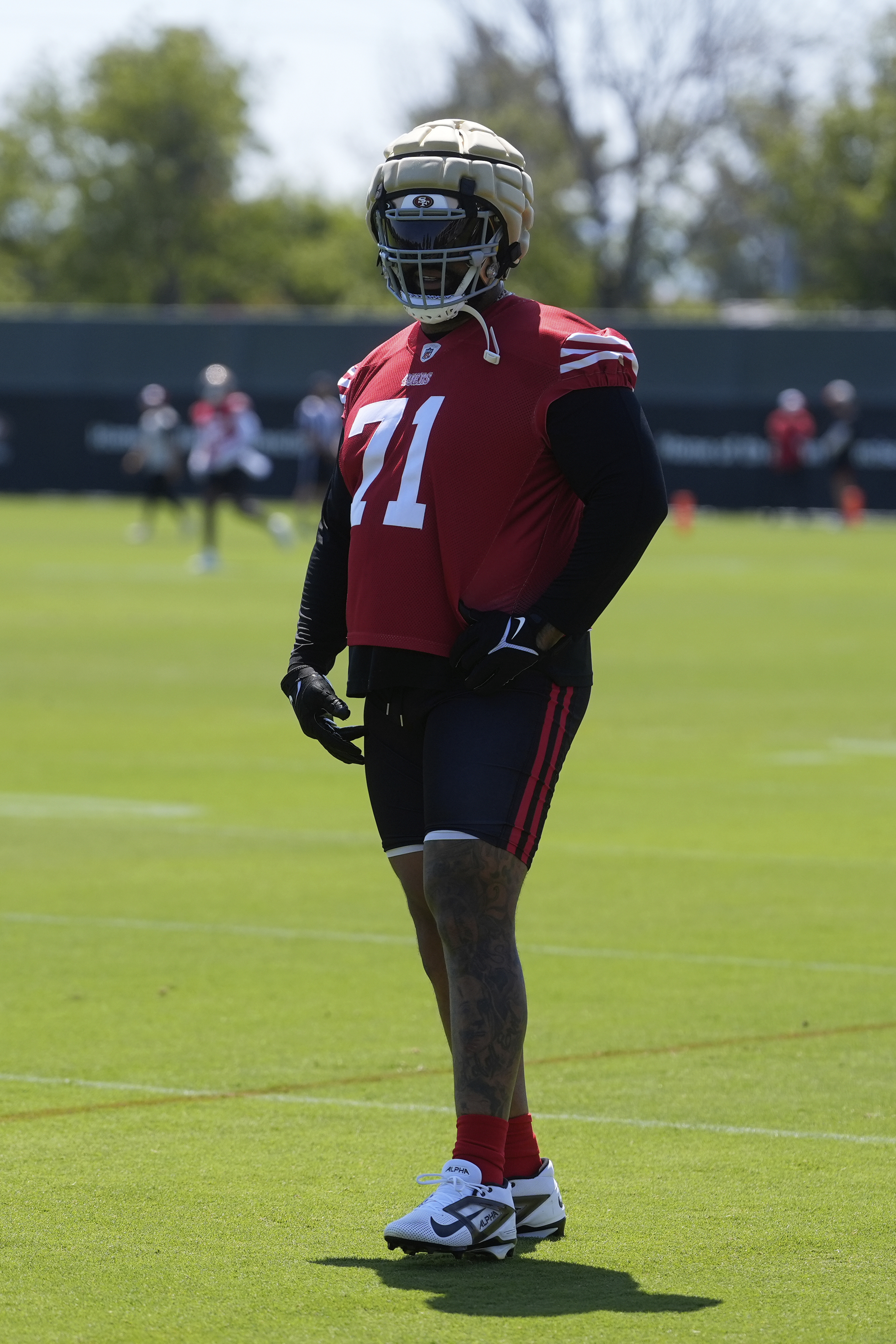 A football player wearing a padded helmet, red jersey with the number 71, and black shorts stands on a grassy field during practice. Trees and others are in the background.