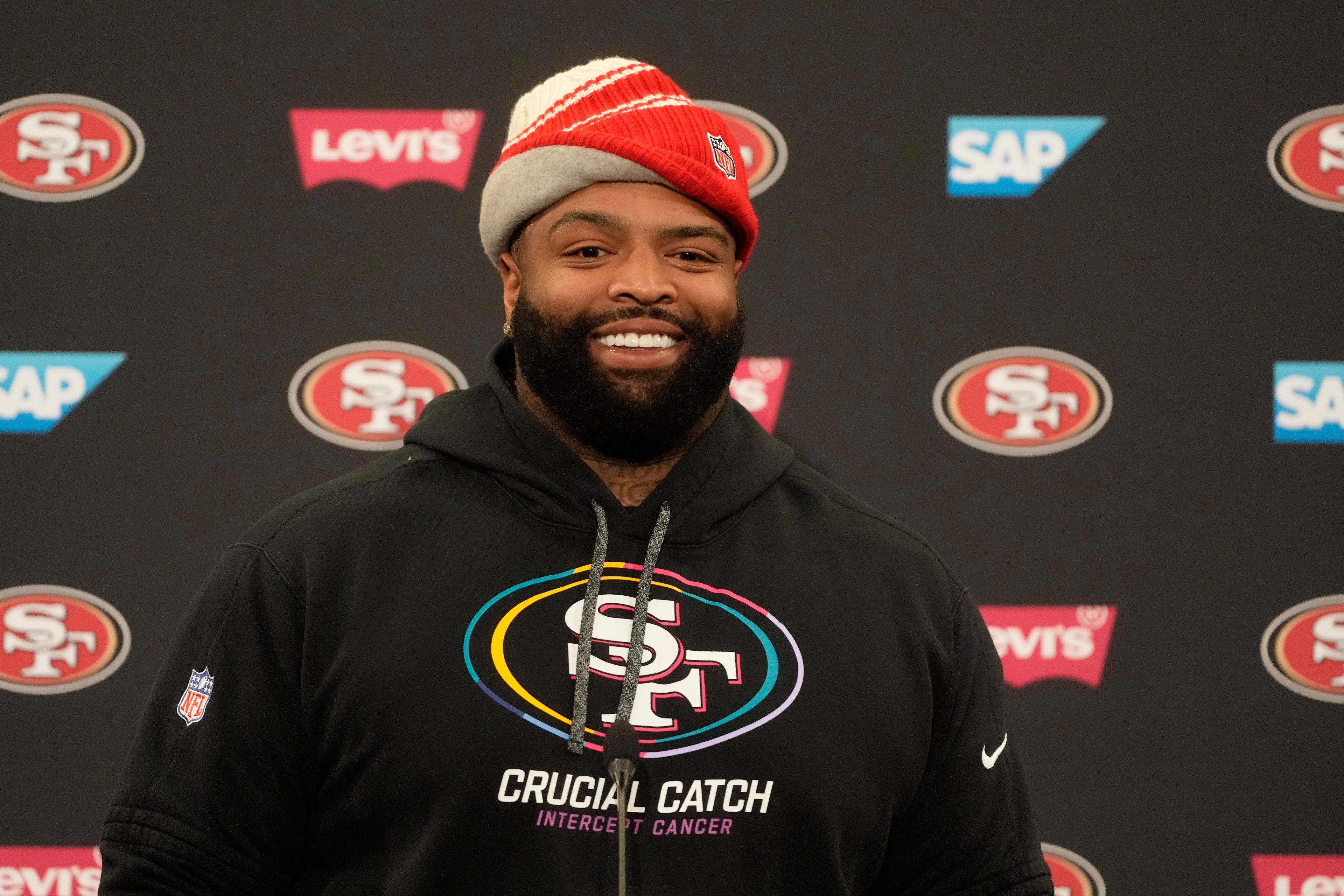 A smiling man in a black hoodie and red beanie stands against a backdrop with 49ers and brand logos. His hoodie reads "Crucial Catch, Intercept Cancer."