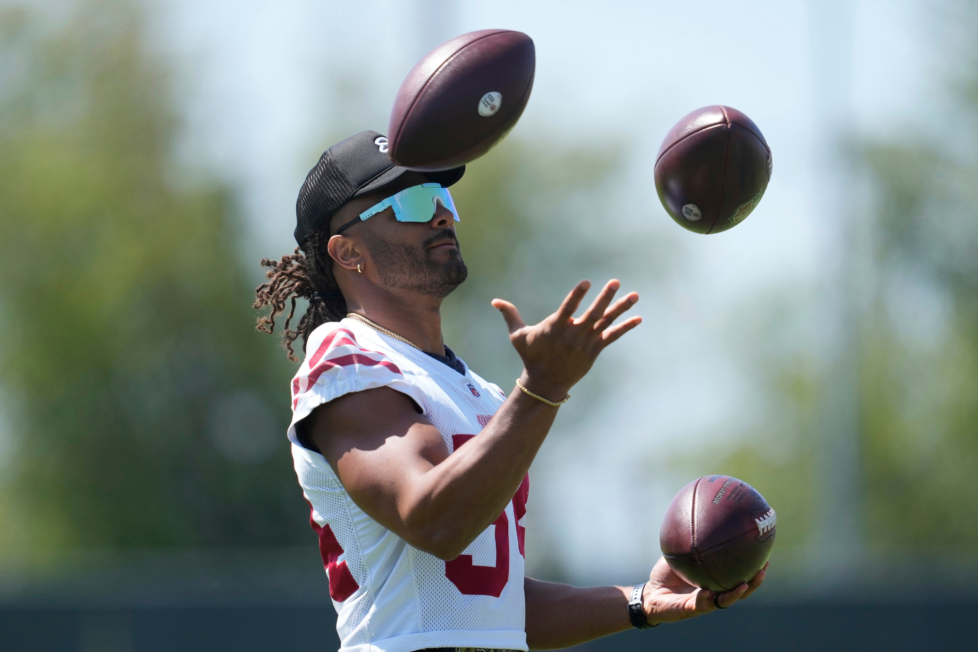 A person wearing a sports jersey, cap, and sunglasses is juggling three American footballs outdoors on a sunny day.