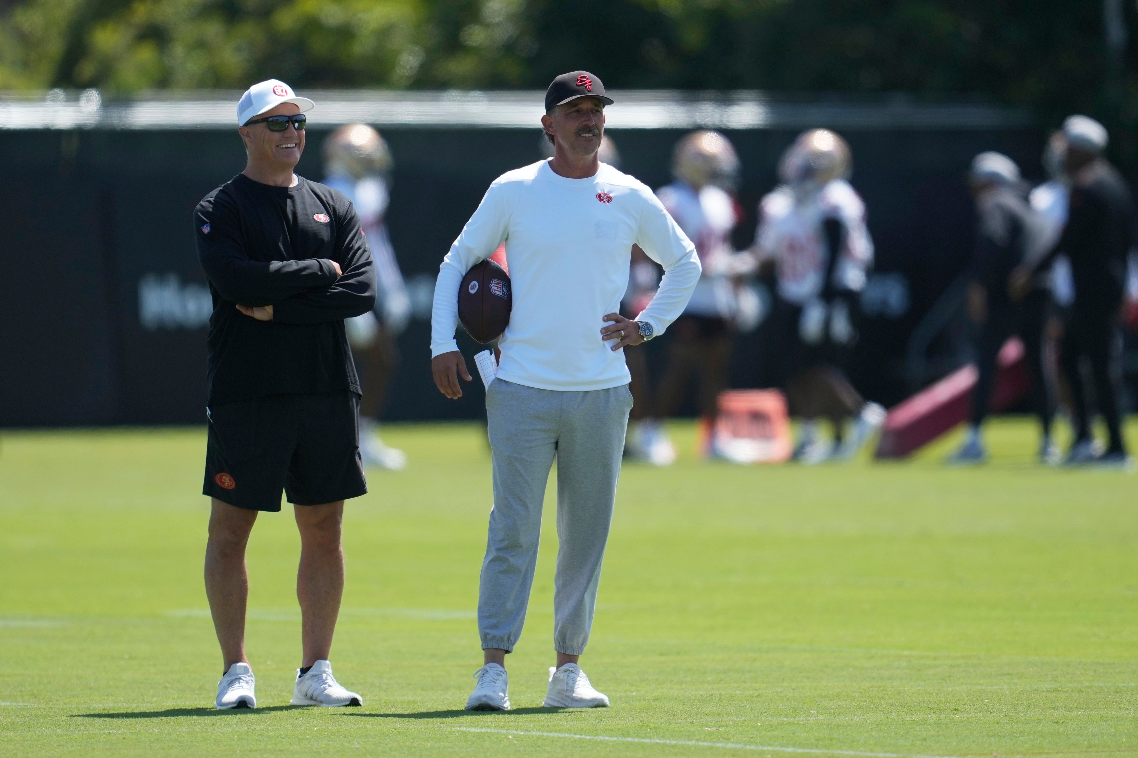 Two men stand on a grassy field during a practice. One wears a black outfit and cap, the other a white top and gray pants, holding a football. Others practice in the background.