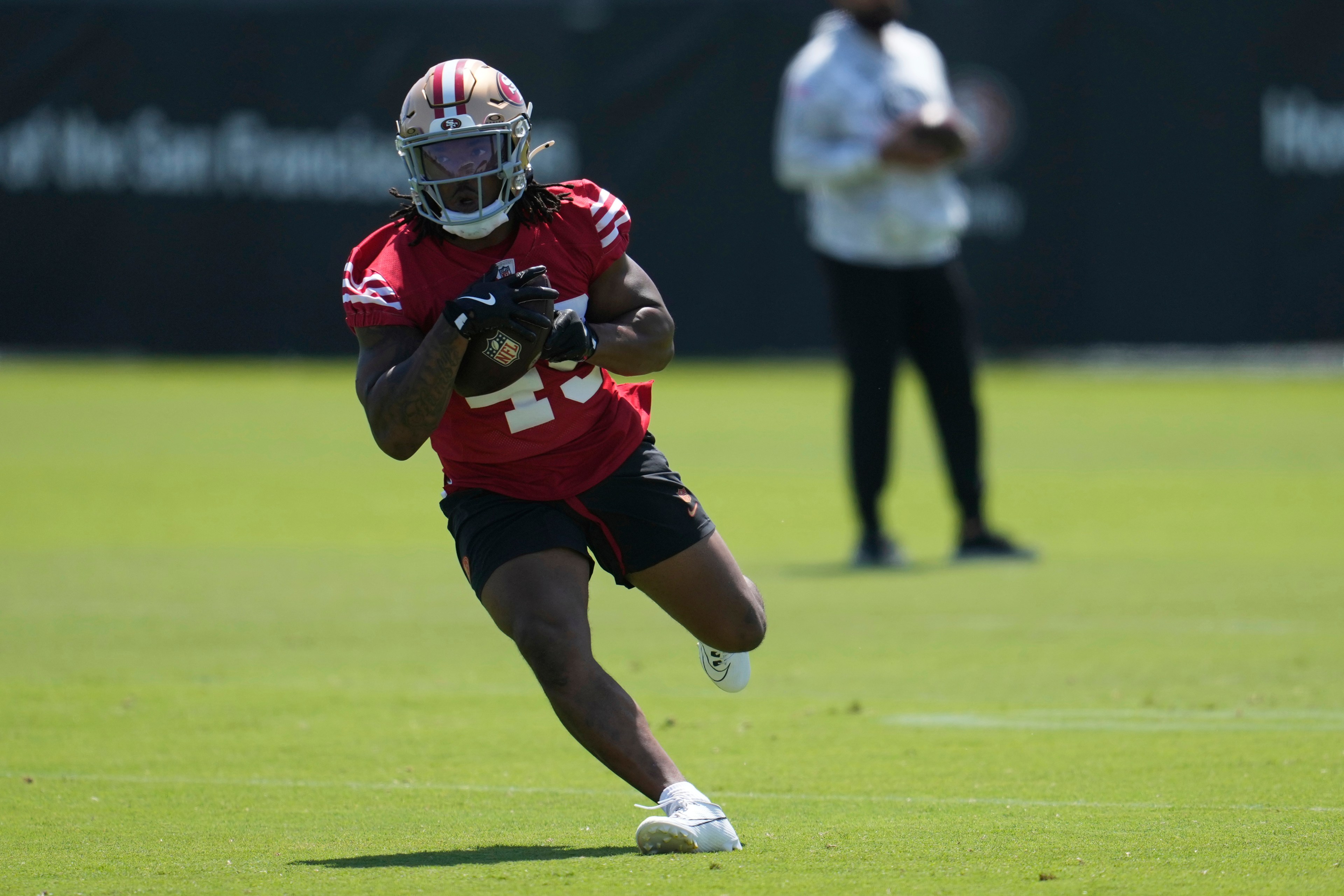 A football player in a red jersey, numbered 23, runs on a grassy field, holding a ball. He's wearing a helmet and gloves, with a person blurred in the background.