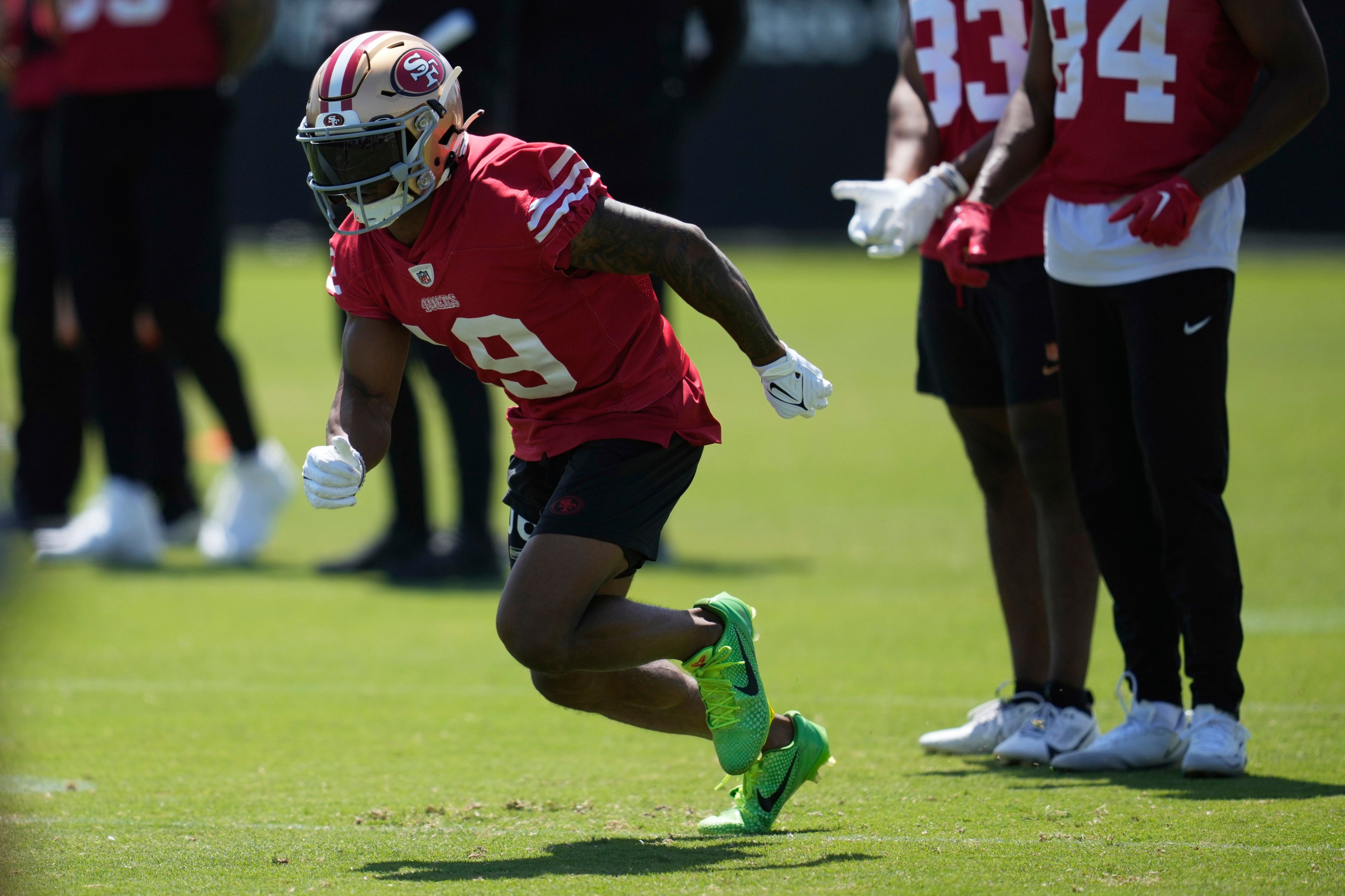 A football player in a red jersey and helmet runs on grass. He wears bright green cleats, and several other players in red jerseys stand nearby.