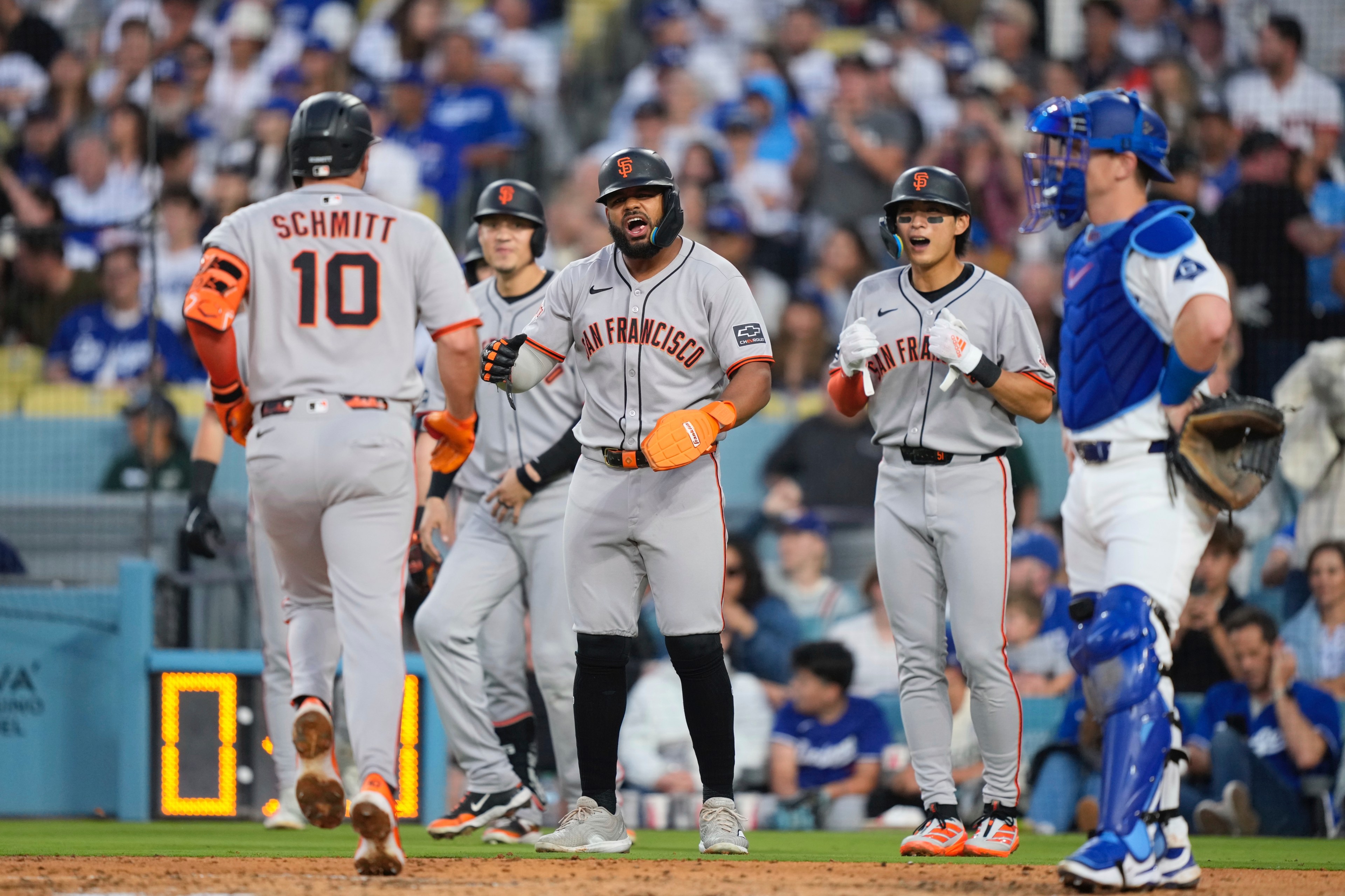Baseball players in gray jerseys with "San Francisco" celebrate on the field, while a player in blue catcher gear stands nearby. The crowd watches in the background.