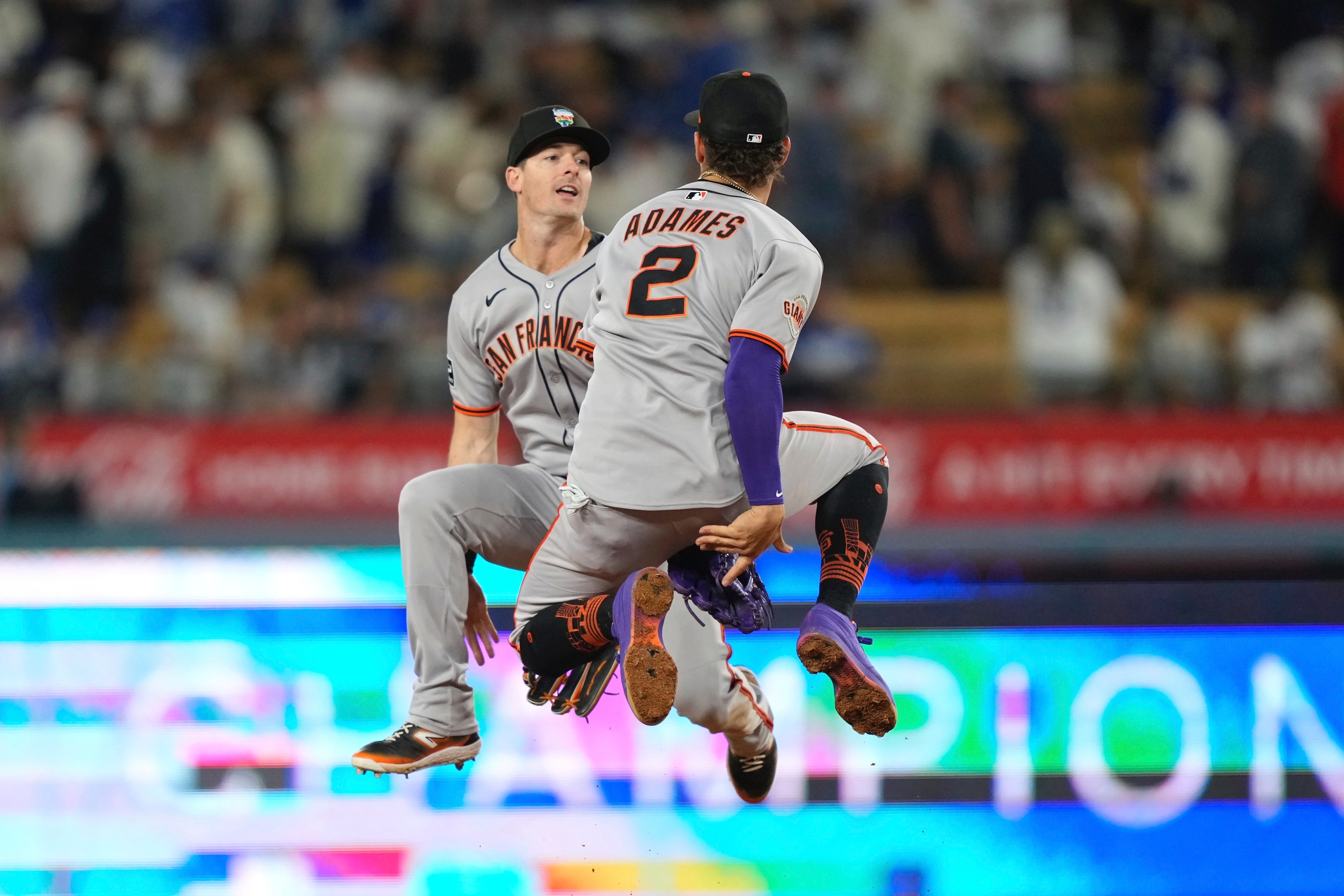 Two baseball players in gray uniforms are mid-air, celebrating. One player's back is visible with &quot;ADAMES&quot; and number 2. They're on a baseball field with a blurred crowd.