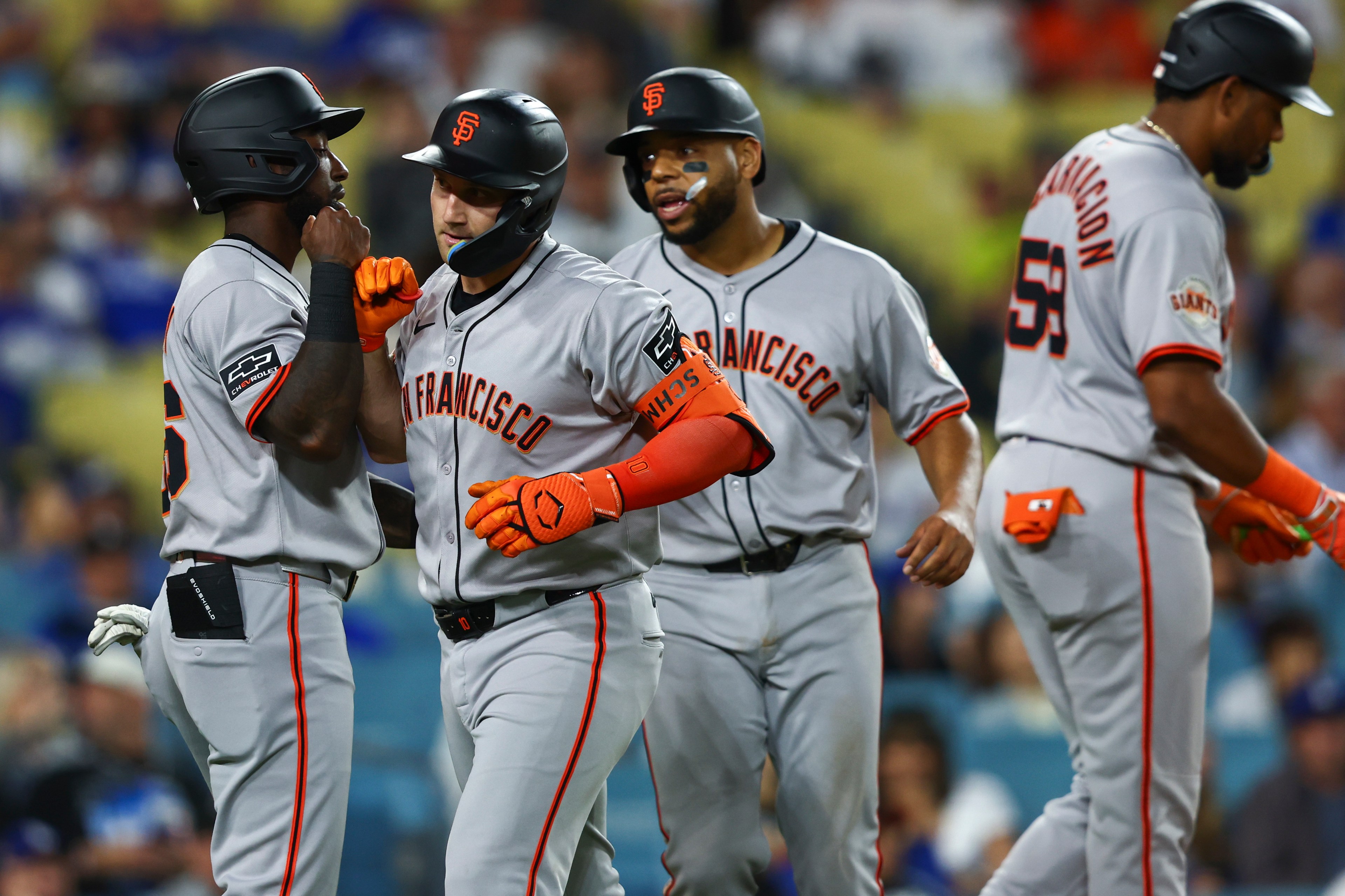Four baseball players in gray &quot;San Francisco&quot; uniforms celebrate on the field, wearing black helmets and orange gloves, with a blurred crowd in the background.