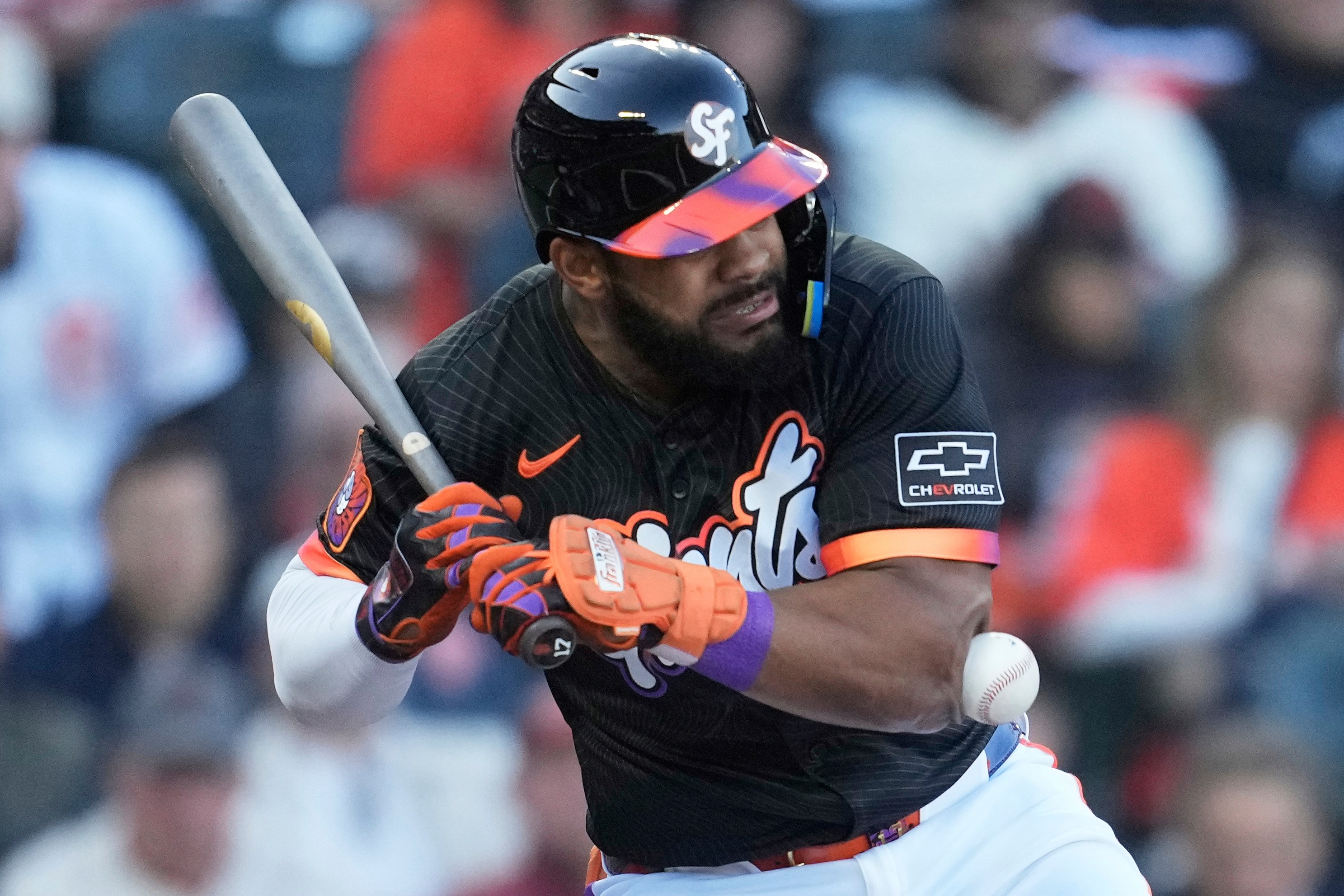 A baseball player in a black uniform with orange and purple accents braces as a ball approaches his chest. He holds a bat and wears a helmet.