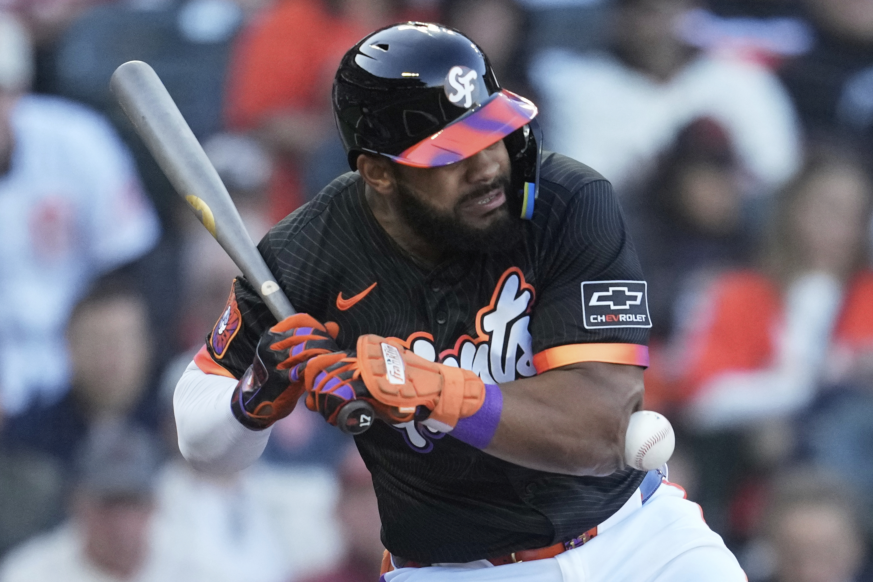 A baseball player in a black uniform with orange and purple accents braces as a ball approaches his chest. He holds a bat and wears a helmet.
