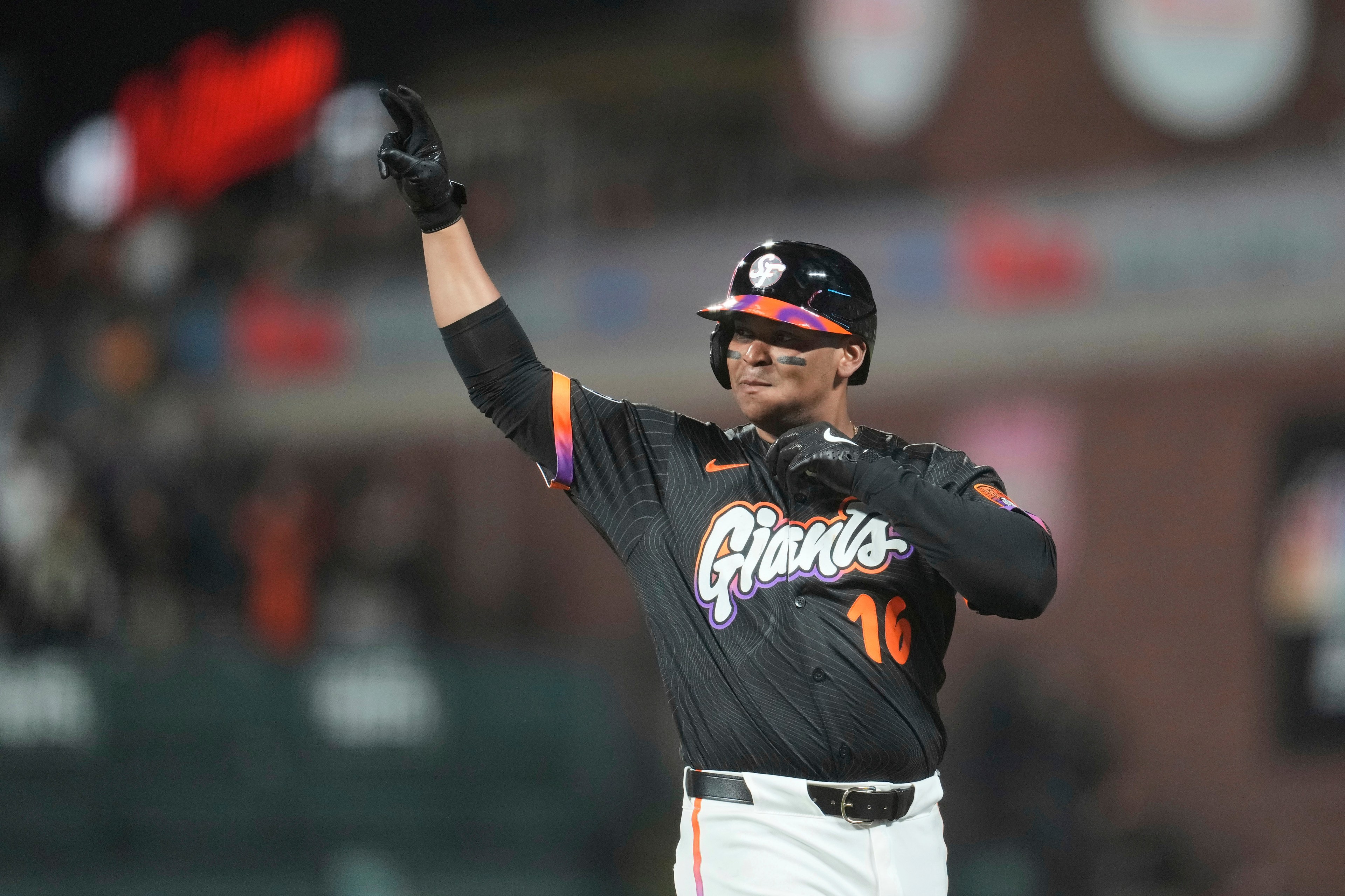 A baseball player in a black "Giants" uniform and helmet is celebrating with his arm raised. He’s wearing black gloves and has face paint under his eyes.