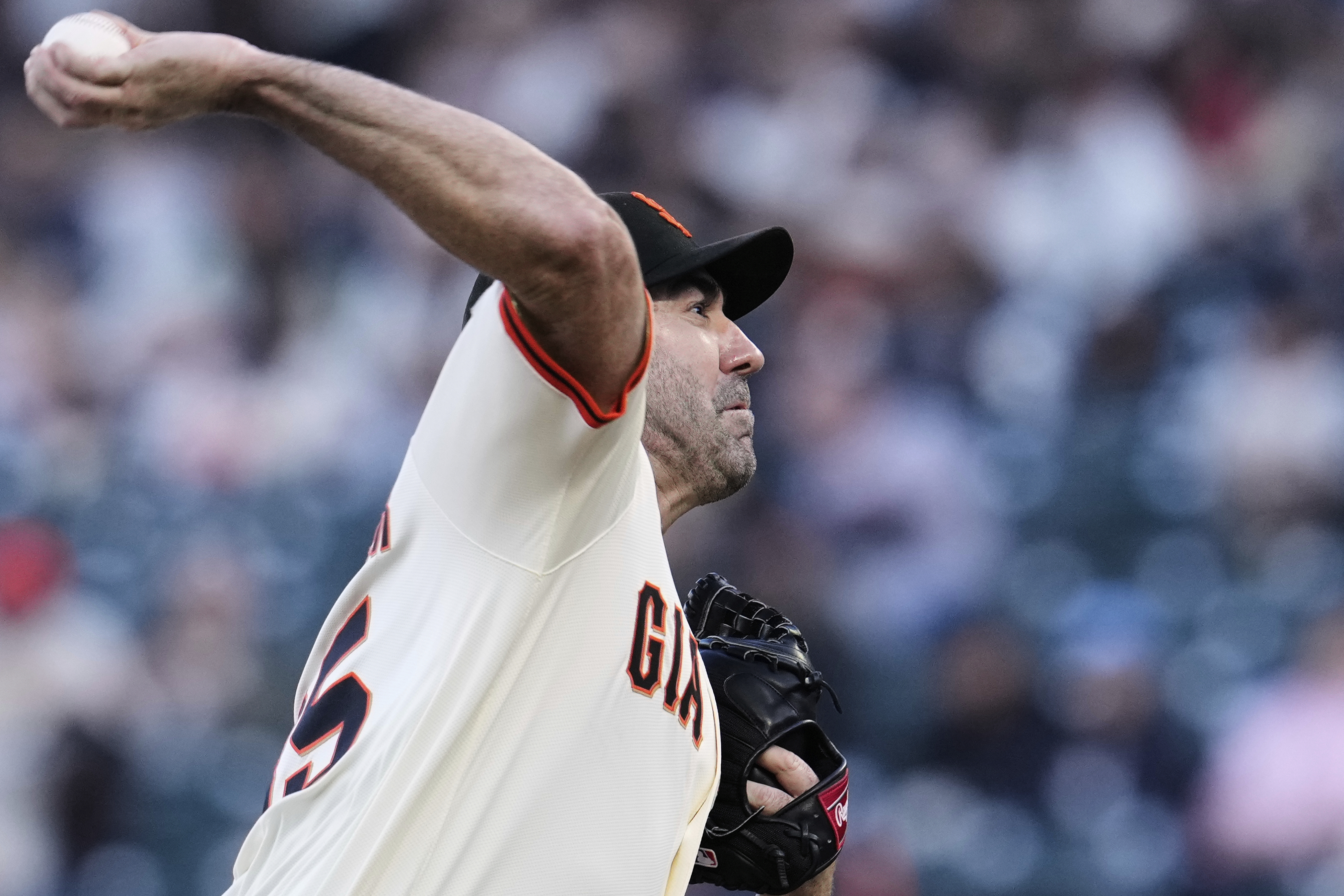 A baseball player in a white jersey with "GI" partially visible is pitching. The background is blurred, focusing on the player's action.