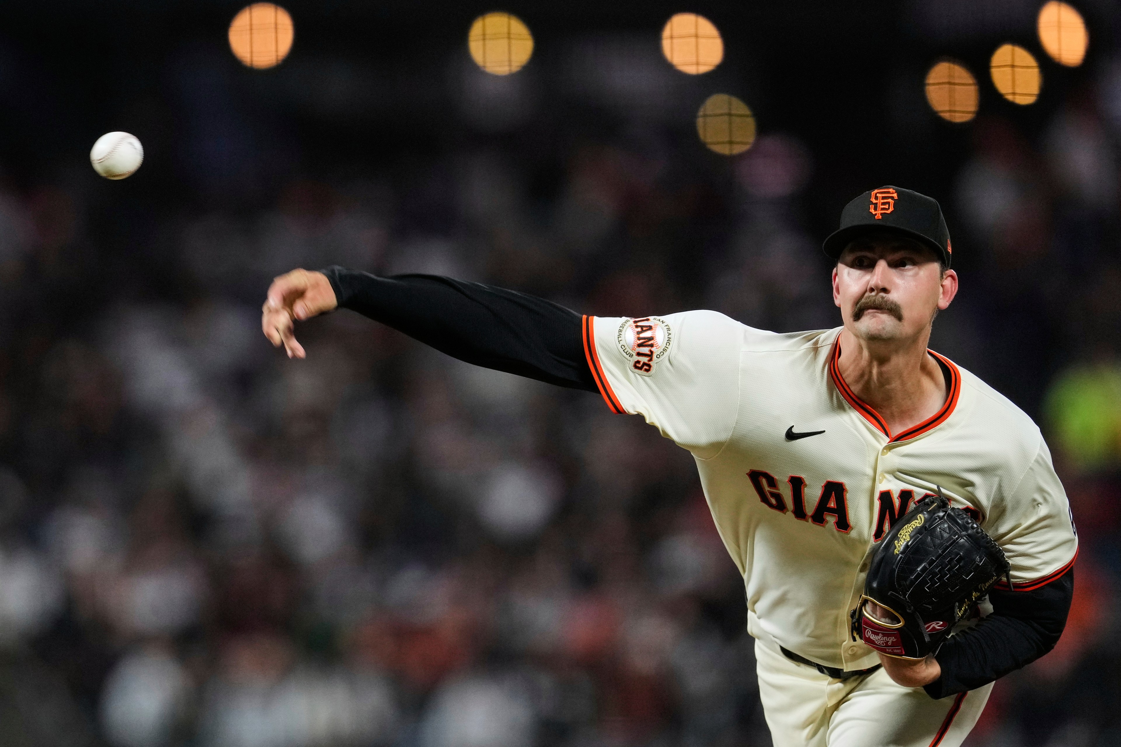 A pitcher in a white Giants uniform with a black glove throws a baseball. The backdrop shows blurred lights and a crowd, creating an action-packed scene.