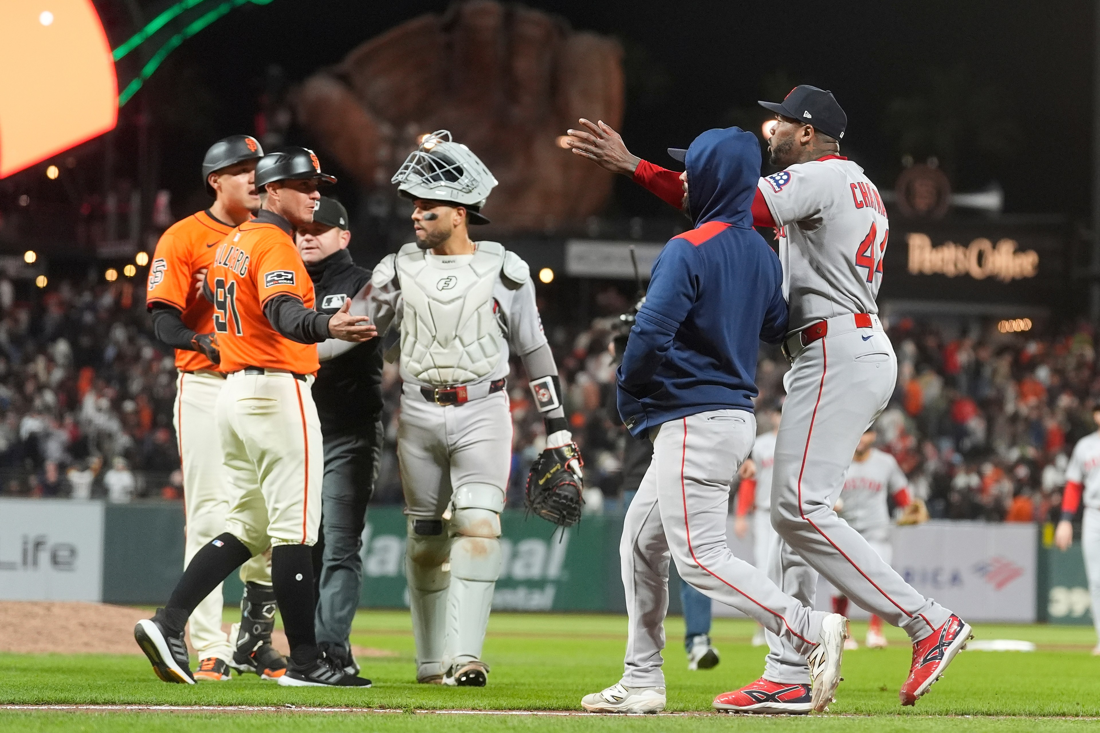 Two baseball teams, one in orange and one in gray, are engaged in a tense exchange on the field with a catcher and umpire intervening.