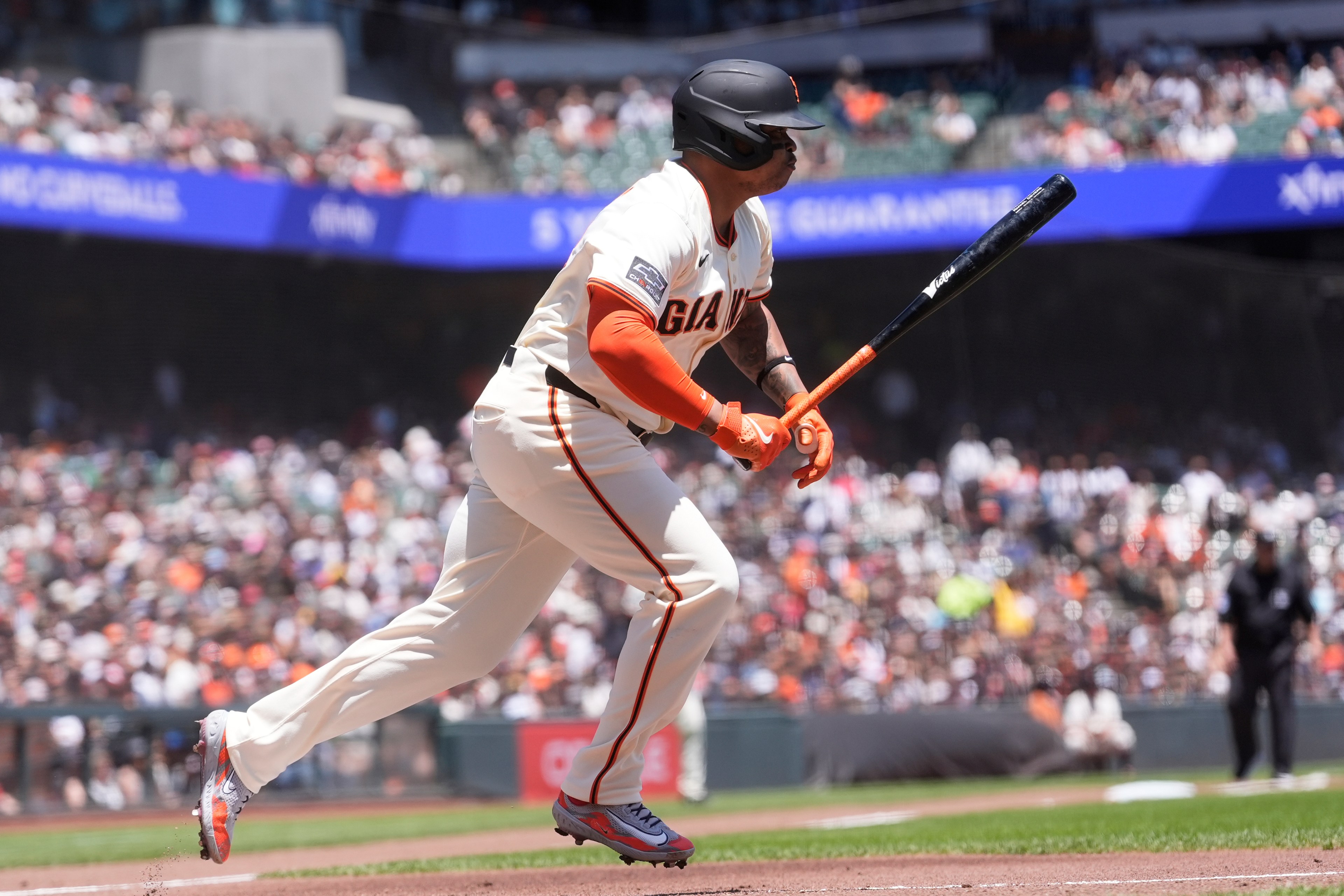 A baseball player in a white uniform and helmet runs with a bat in hand during a game. The background shows a blurred crowd under a blue advertisement banner.