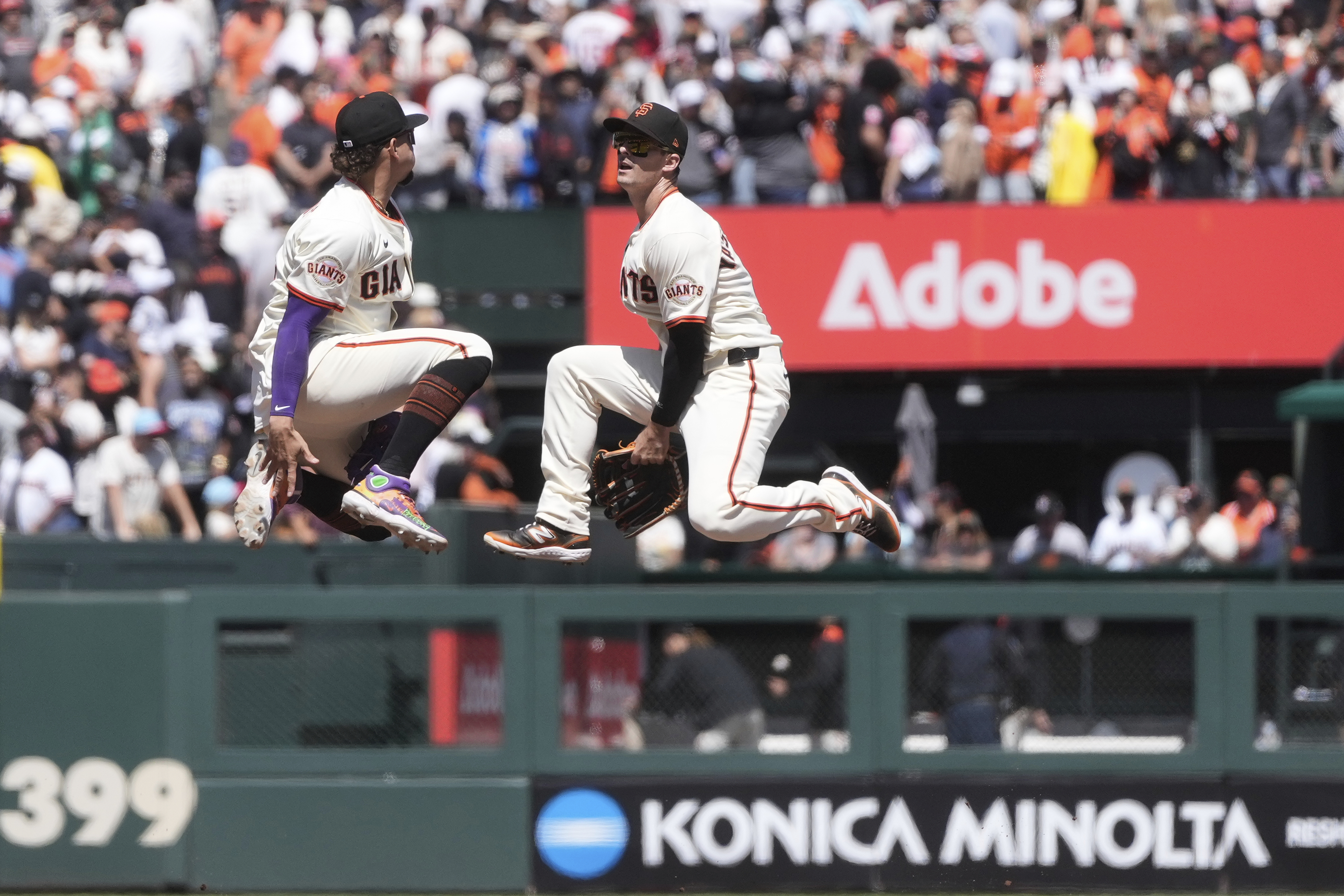 Two baseball players from the Giants, in mid-air, celebrate during a game. The crowd watches in the background, with advertisements visible around.