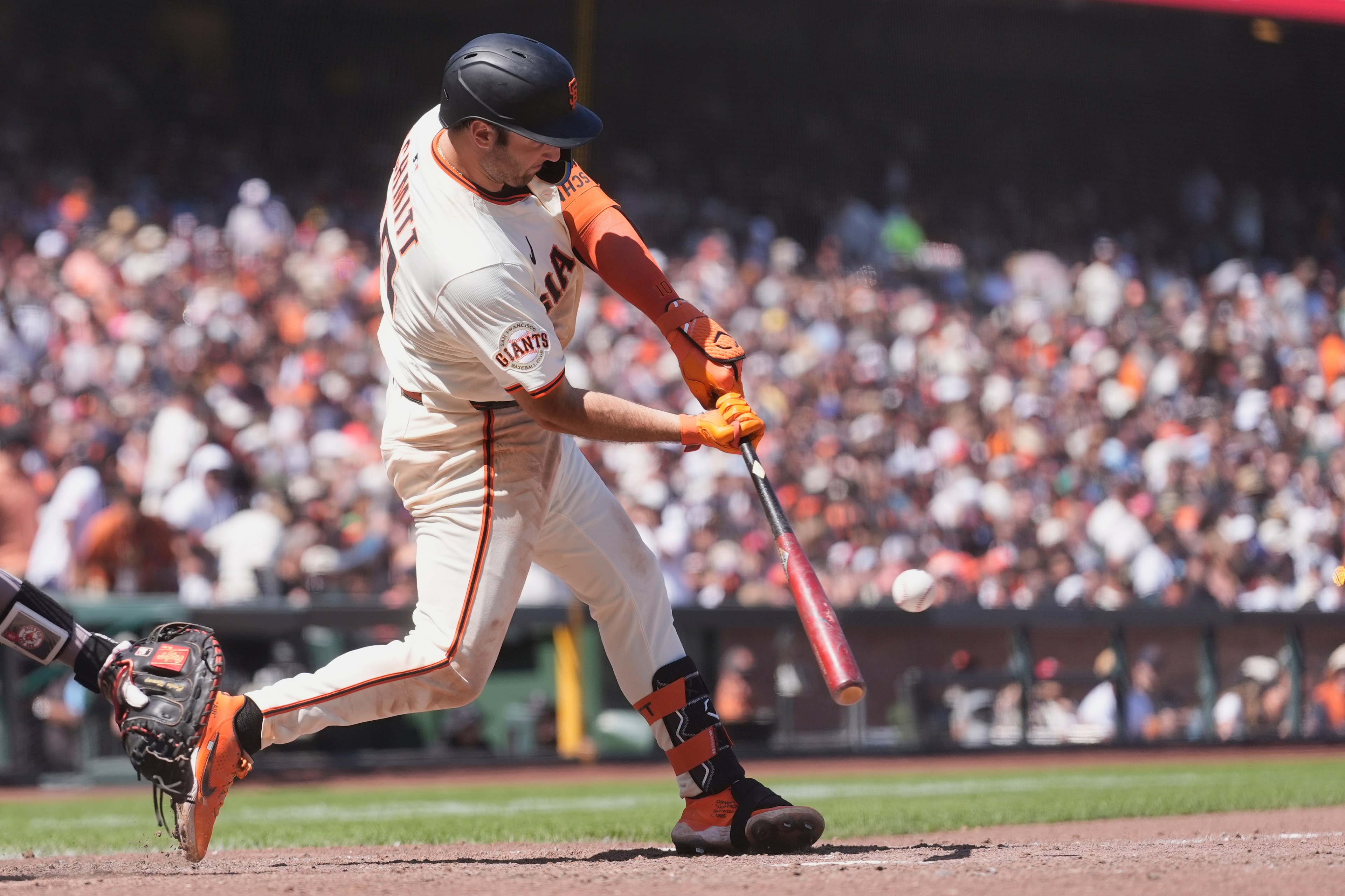 A baseball player in a white uniform is swinging a bat at a ball. The crowd in the stands is blurred in the background, and the player wears a helmet and orange gloves.