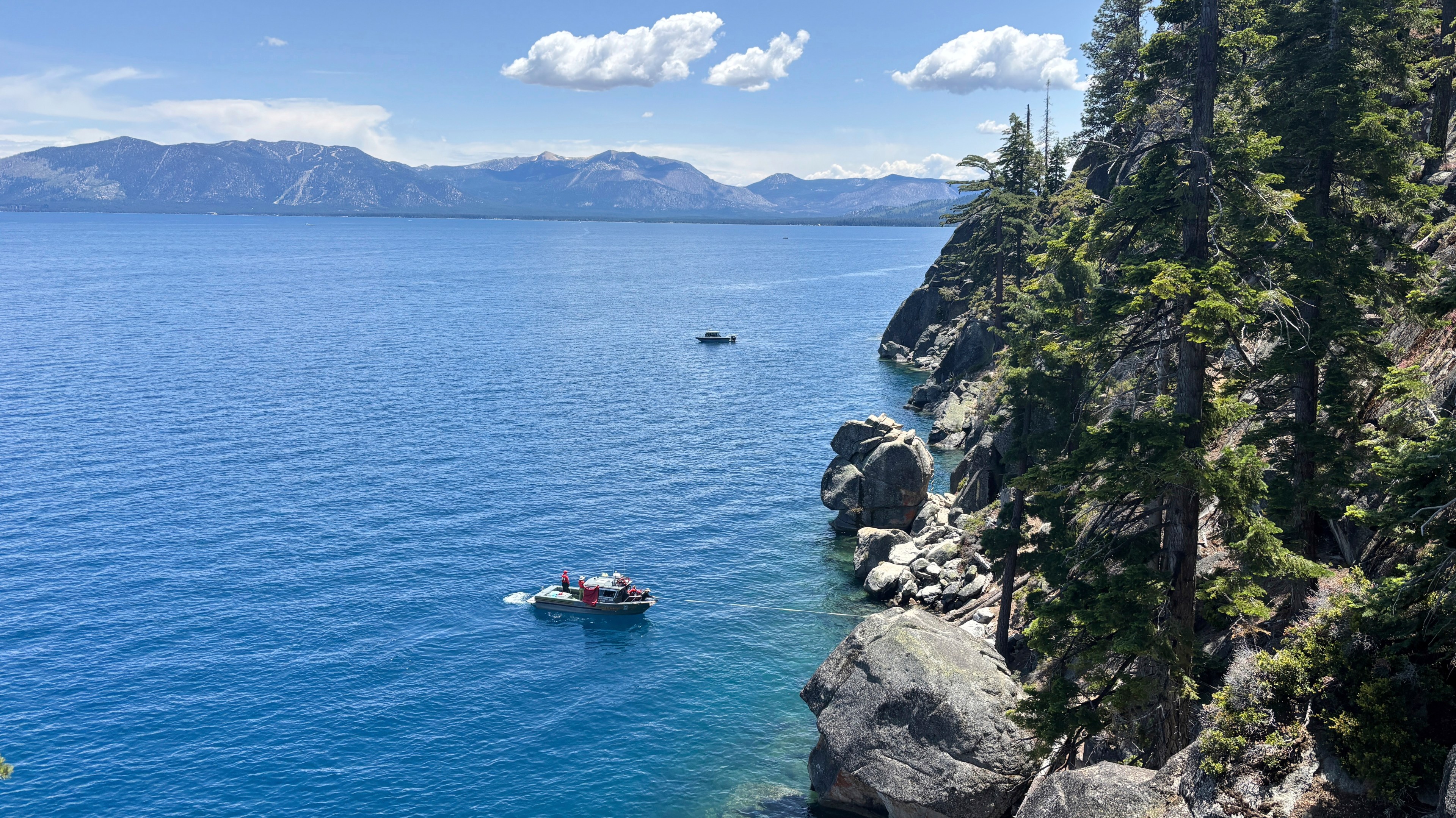 A vast lake bordered by rocky cliffs and tall pine trees under a clear blue sky. Boats are visible on the water, with mountains in the distance.
