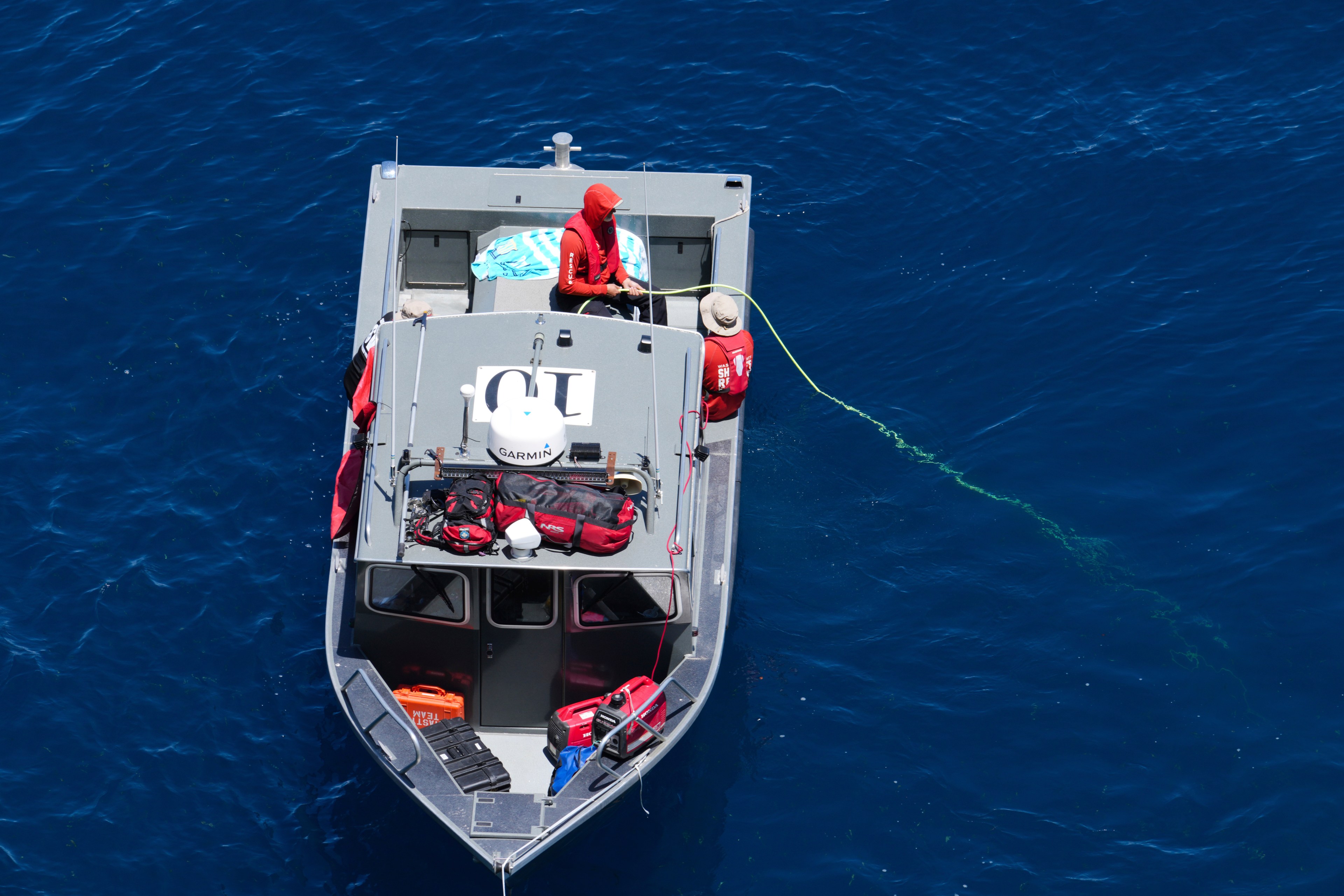 Two people in red clothing are on a small gray boat in deep blue water. One person holds a green rope extending into the water. Onto the boat's roof, various gear is secured.