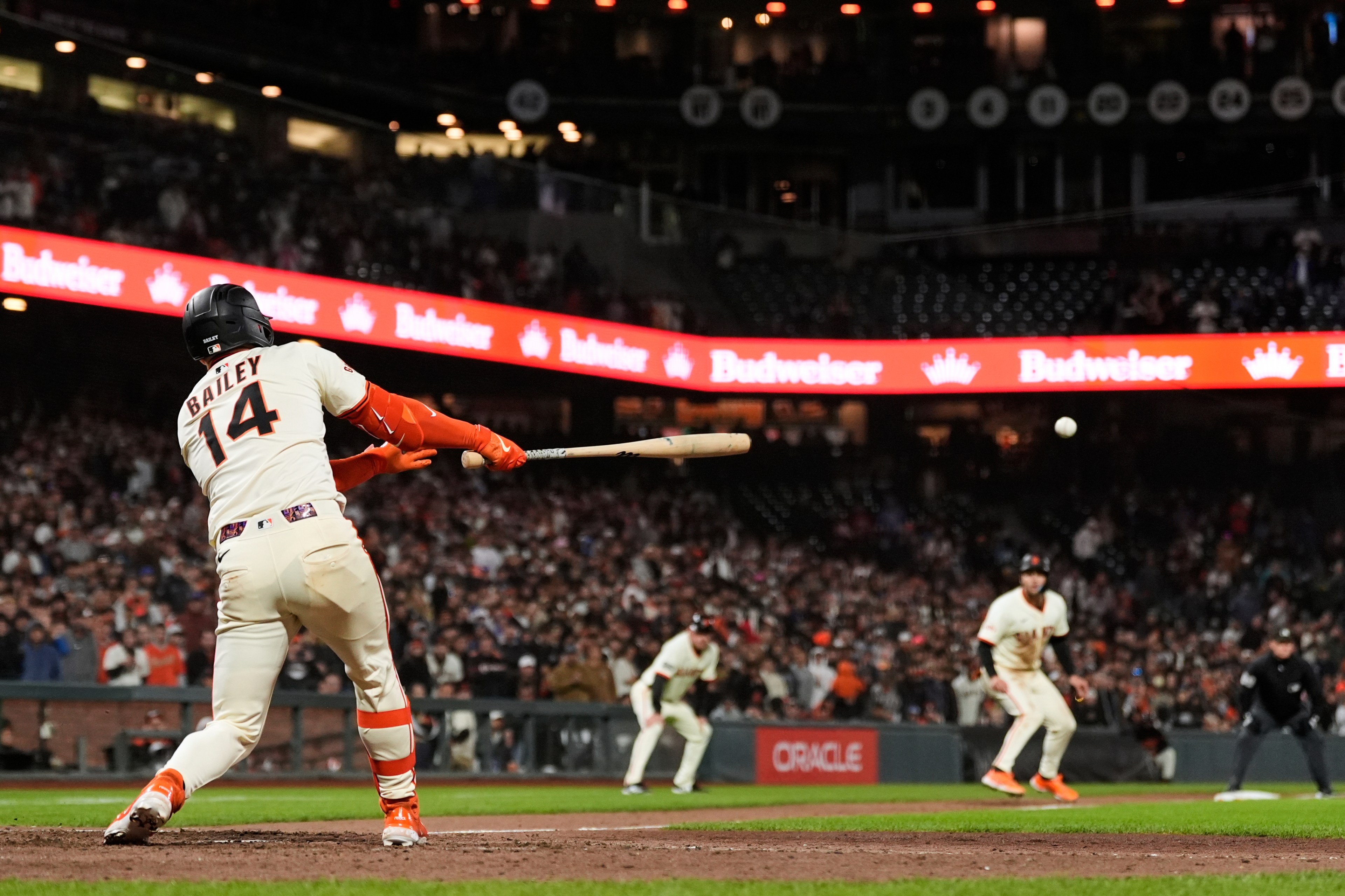 A baseball player in a white uniform with orange accents swings a bat in a stadium. The crowd is blurred in the background, illuminated by advertising lights.