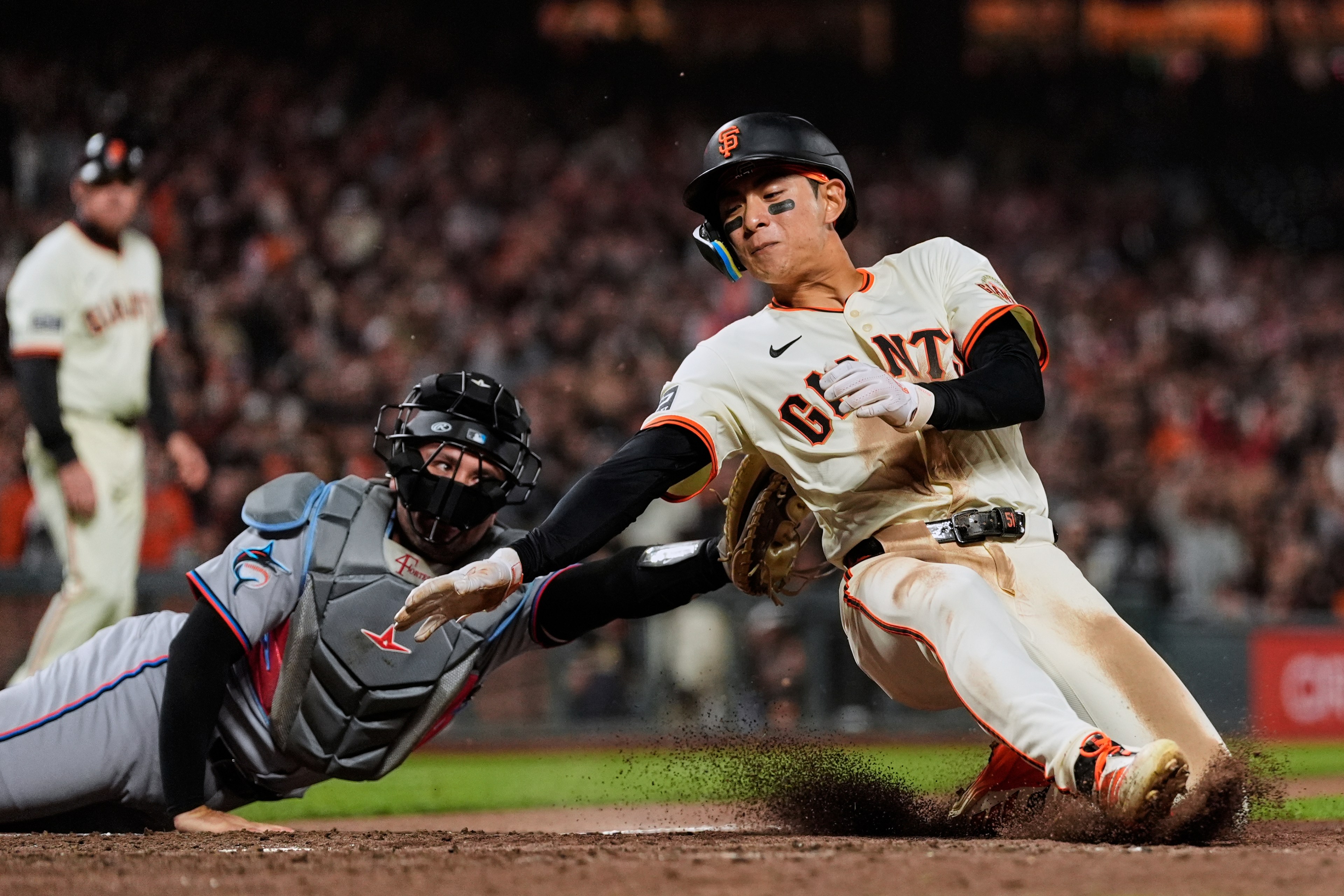 A baseball player in a white "Giants" jersey slides into home base as a catcher in grey gear tries to tag him out. Dirt is flying in the air.