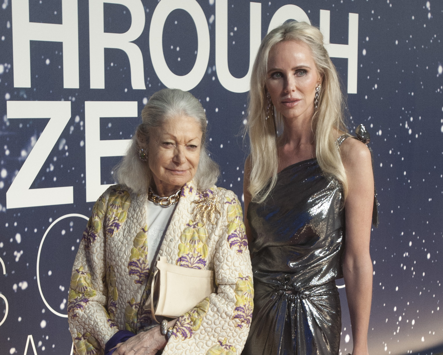 Two women stand together; one wears a quilted floral jacket, the other a shiny silver dress. They pose in front of a backdrop with large text and a starry design.