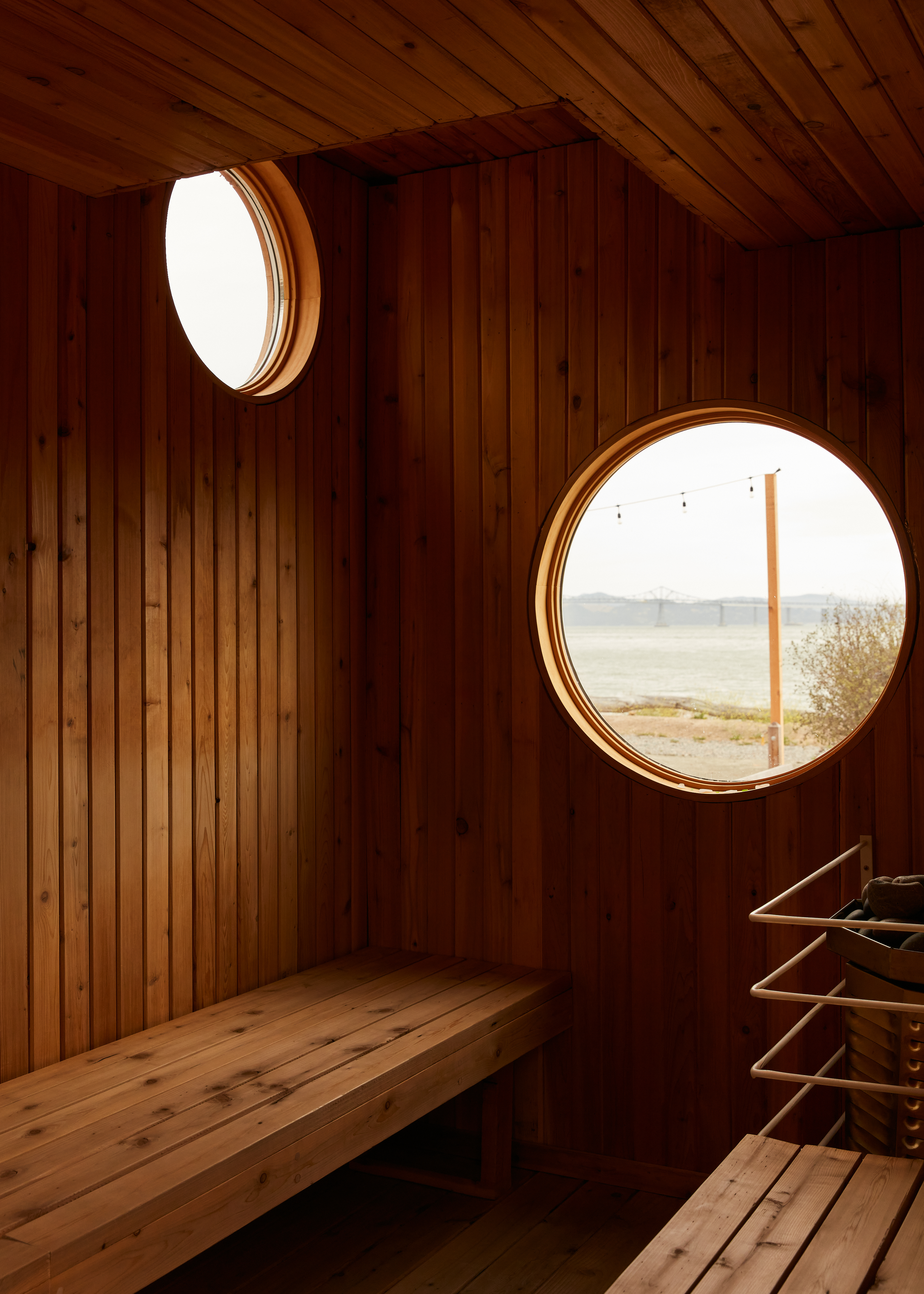 A wooden sauna features circular windows, one large with an outdoor view and a smaller one above. Benches line the walls, and a heater with racks is in the corner.