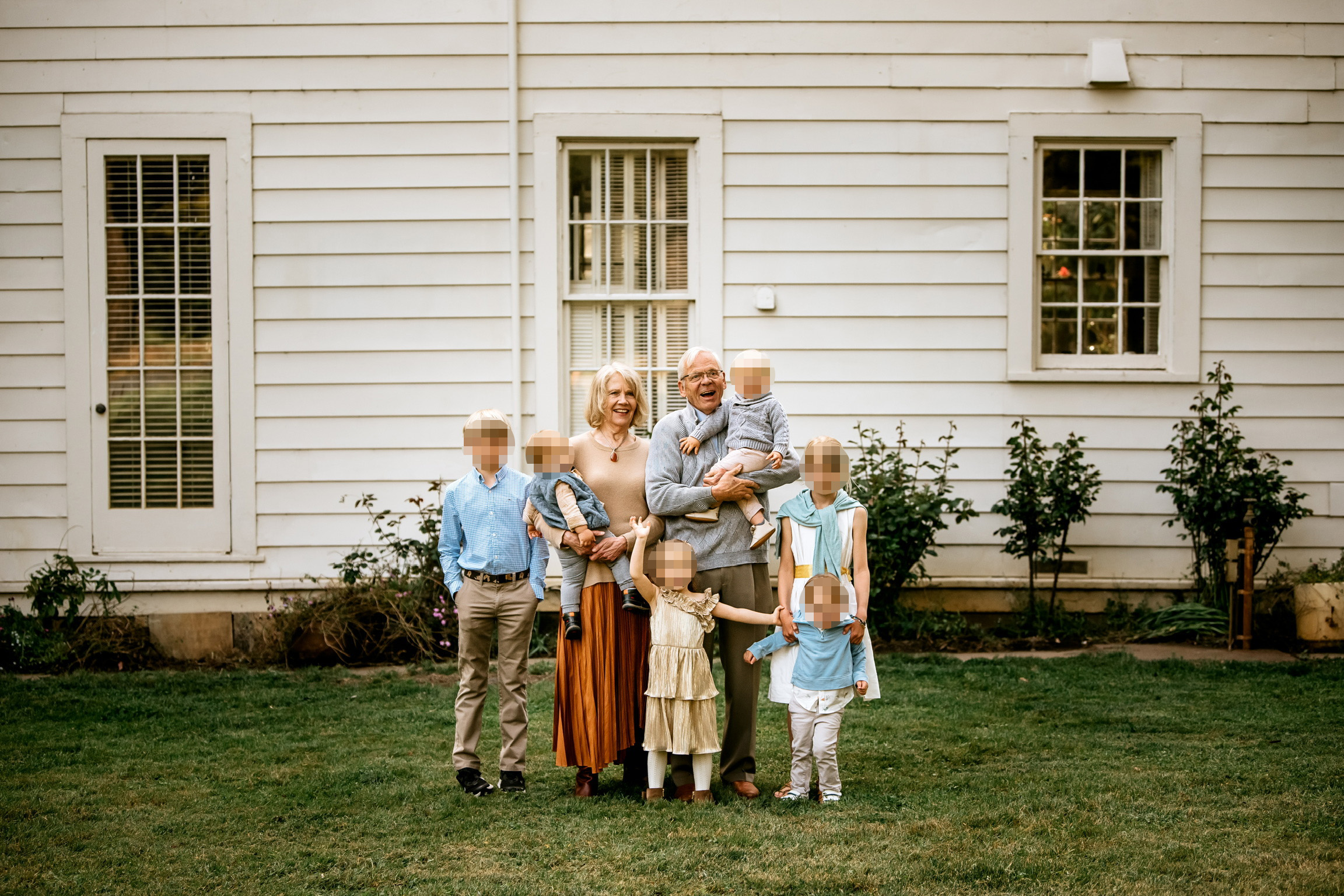 A group of seven people stands in front of a white house. An older couple is in the center, surrounded by children, with greenery and two windows in the background.