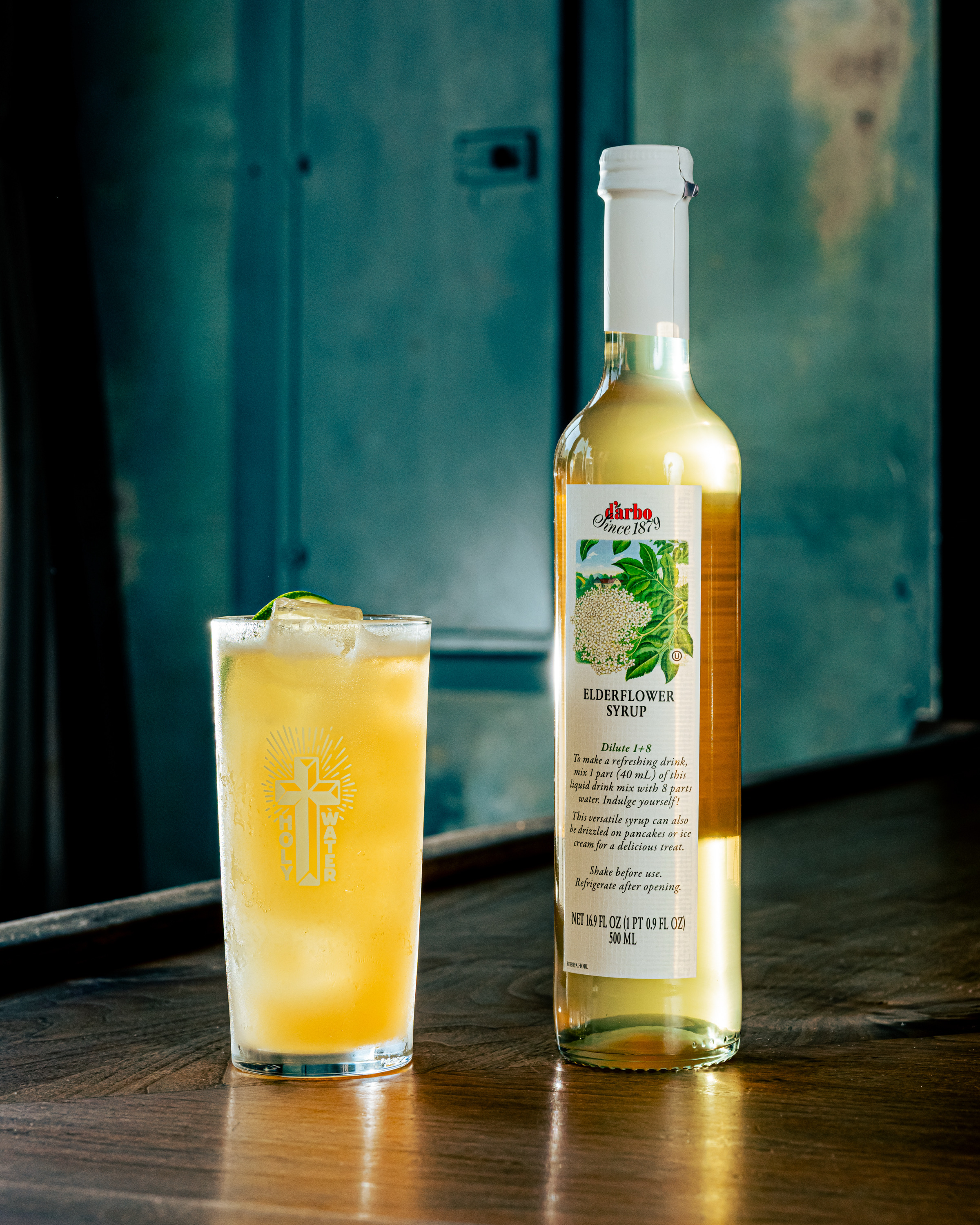 A tall glass filled with an iced, orange drink sits beside a bottle of elderflower syrup. The wooden surface reflects a dimly lit, industrial background.