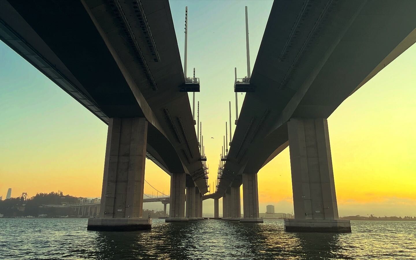 The image shows the underside of a bridge at sunset, with a vibrant orange-yellow sky. The bridge's massive pillars rise from the water, creating a symmetrical view.